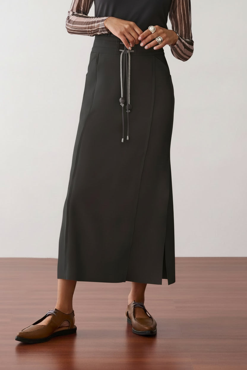 Modelo veste uma saia midi preta que apresenta fenda lateral, cordão bicolor com ponteiras, recortes nas laterais e sapatos marrons de bico fino.