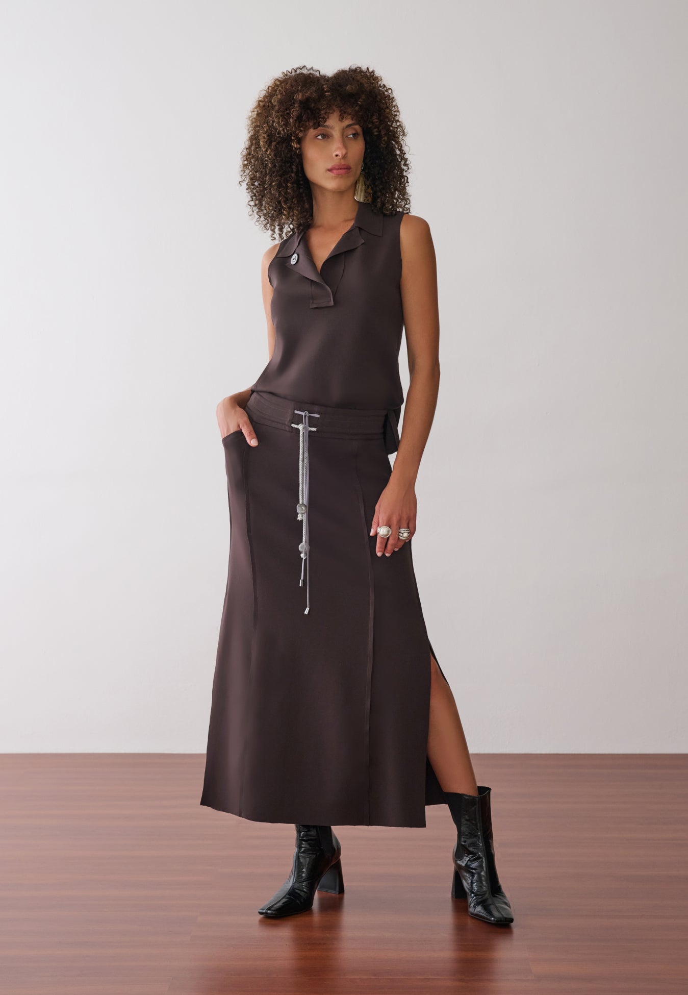Modelo veste uma blusa marrom sem mangas com gola polo aberta, combinada com uma saia midi de mesma cor que apresenta fenda lateral, cordão bicolor com ponteiras, recortes nas laterais e botas pretas de verniz.