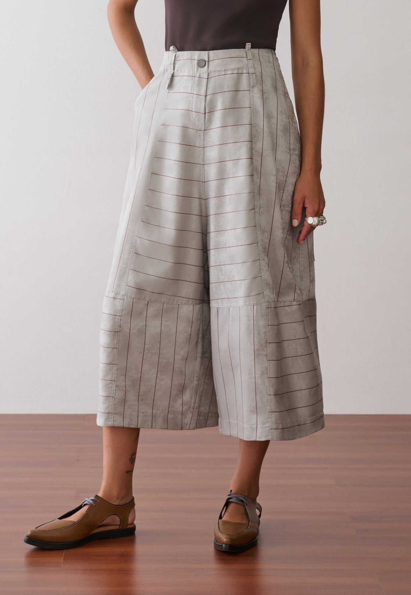 Modelo veste uma regata marrom, combinada com calças pantacourt de cintura alta cinza com listras marrons, bolsos laterais e sapatos marrons de bico fino.