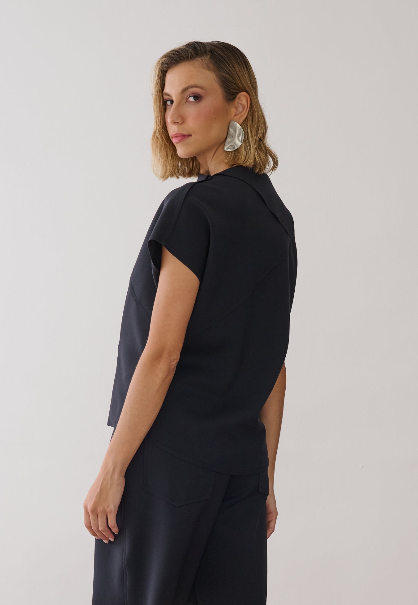 Modelo de costas veste uma blusa preta de gola alta e mangas curtas com costuras geométricas, combinada com bermuda de alfaiataria na mesma cor.