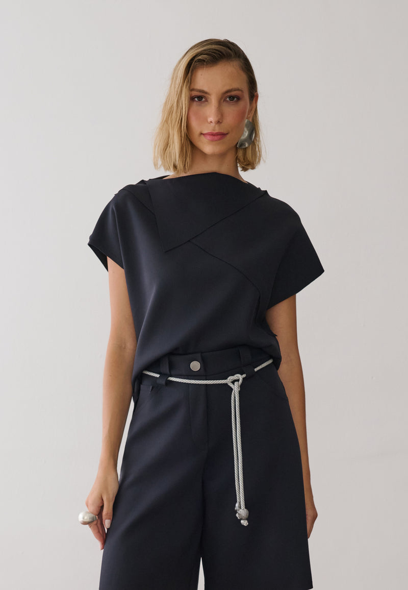 Modelo veste uma blusa preta de gola alta e mangas curtas com costuras geométricas, combinada com bermuda de alfaiataria na mesma cor, cordão off white na cintura.