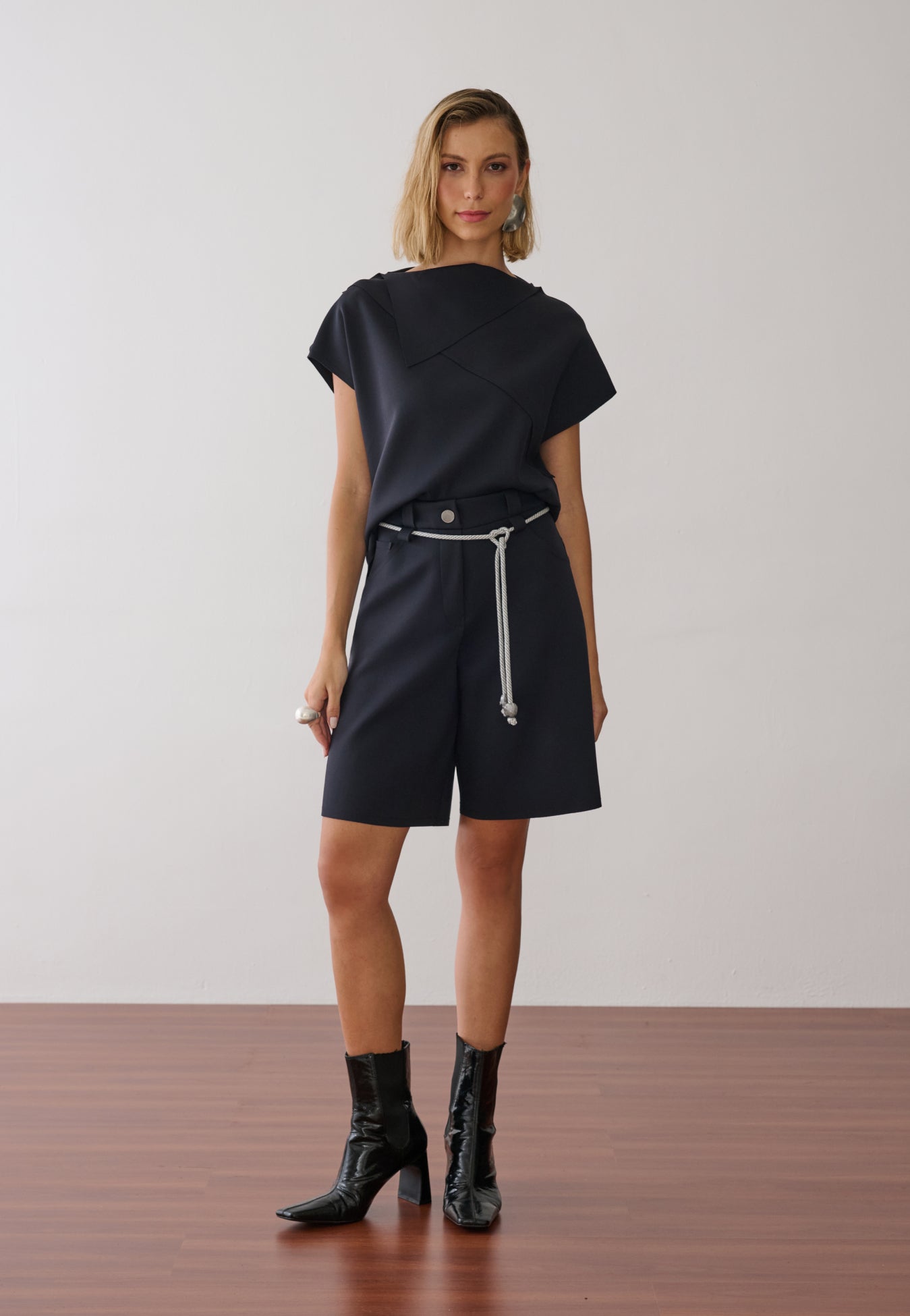 Modelo veste uma blusa preta de gola alta e mangas curtas com costuras geométricas, combinada com bermuda de alfaiataria na mesma cor, cordão off white na cintura e botas pretas de verniz.