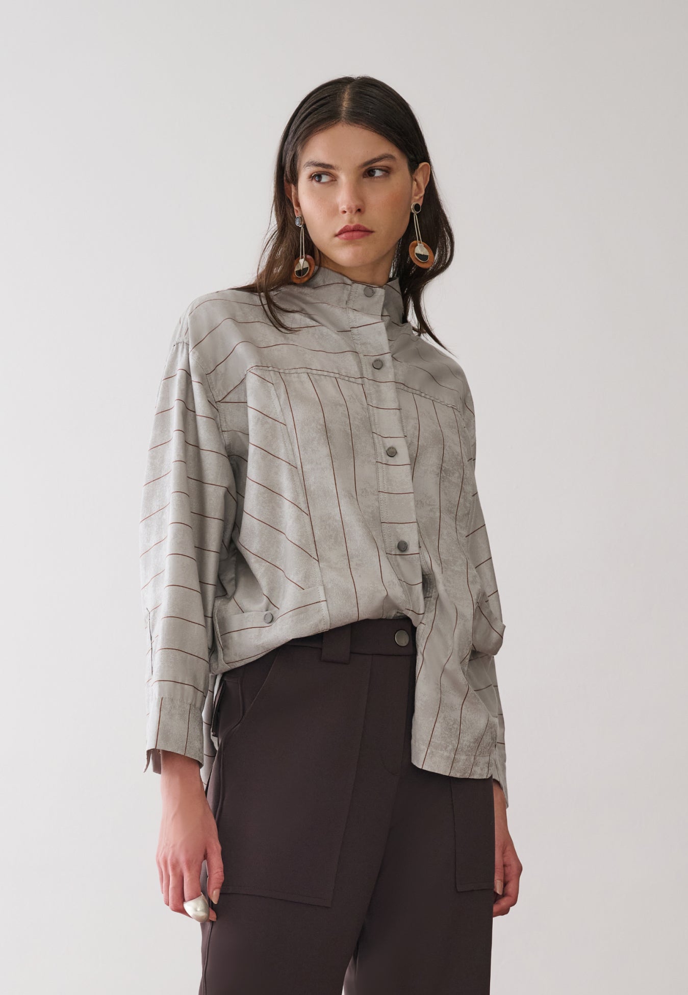 Modelo veste uma camisa cinza de gola alta com padronagem de listras marrons e abotoamento frontal, combinada com calças marrons de bolsos utilitários amplos.