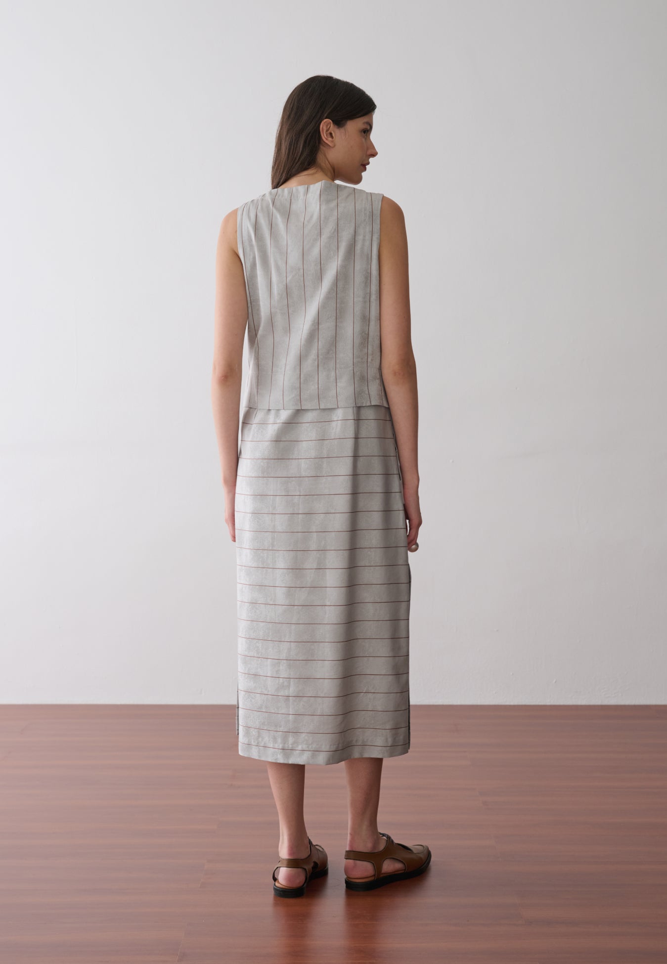Modelo de costas exibe um vestido midi cinza com listras marrons sem mangas, recorte horizontal abaixo da cintura e sapatos marrons de bico fino.