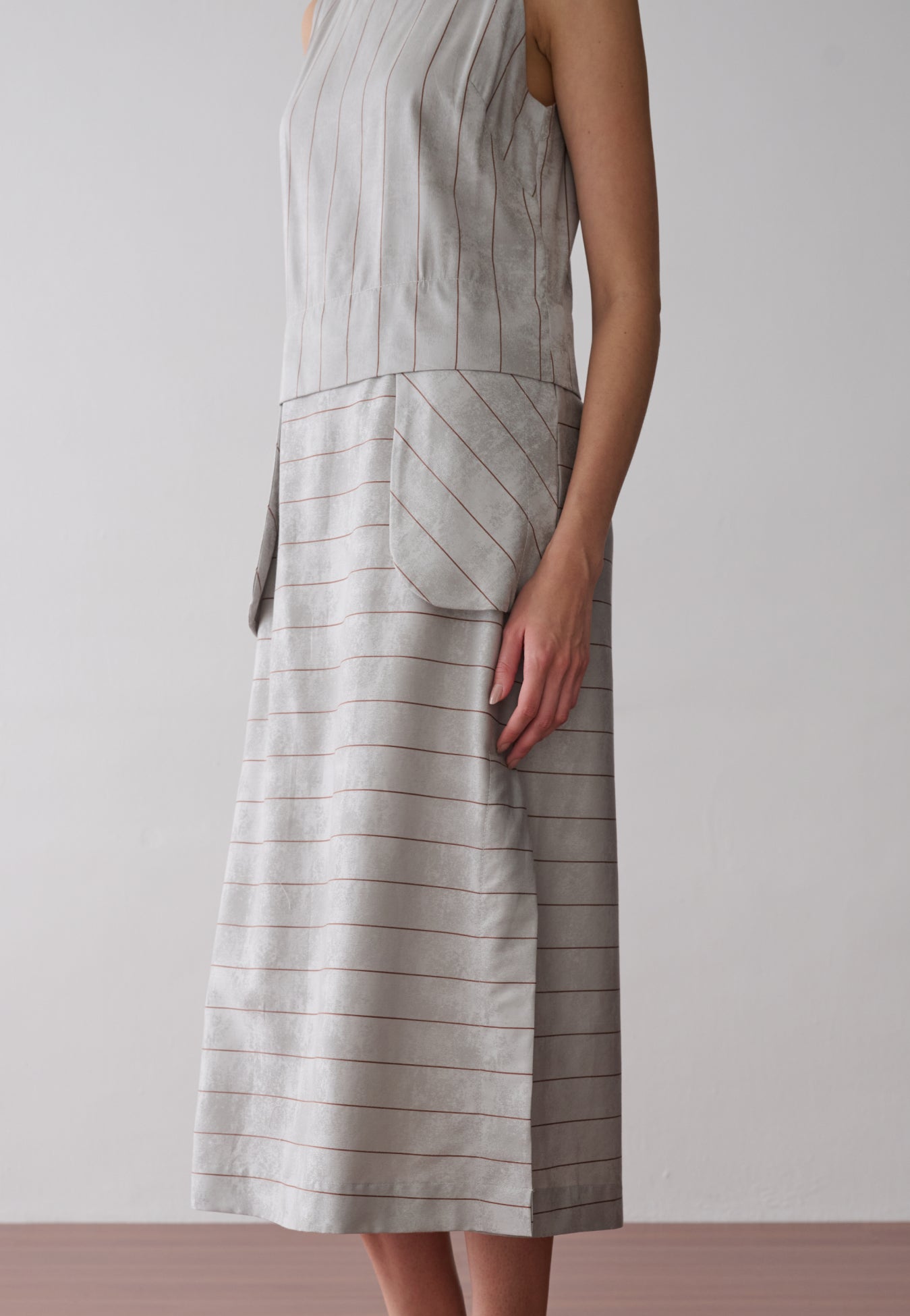 Modelo usa um vestido midi cinza com listras marrons sem mangas, apresentando bolsos frontais arredondados.