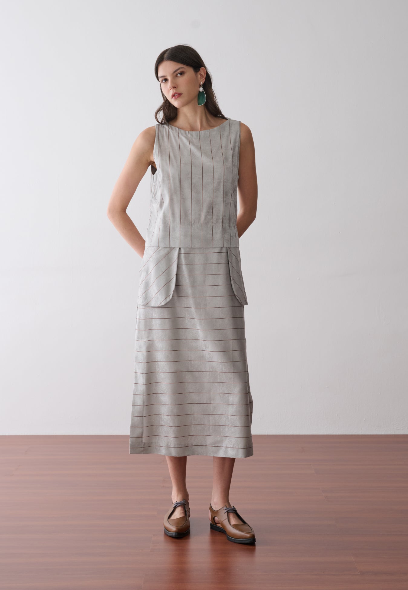 Modelo veste um vestido midi cinza com listras marrons sem mangas, apresentando bolsos frontais arredondados e sapatos marrons de bico fino.