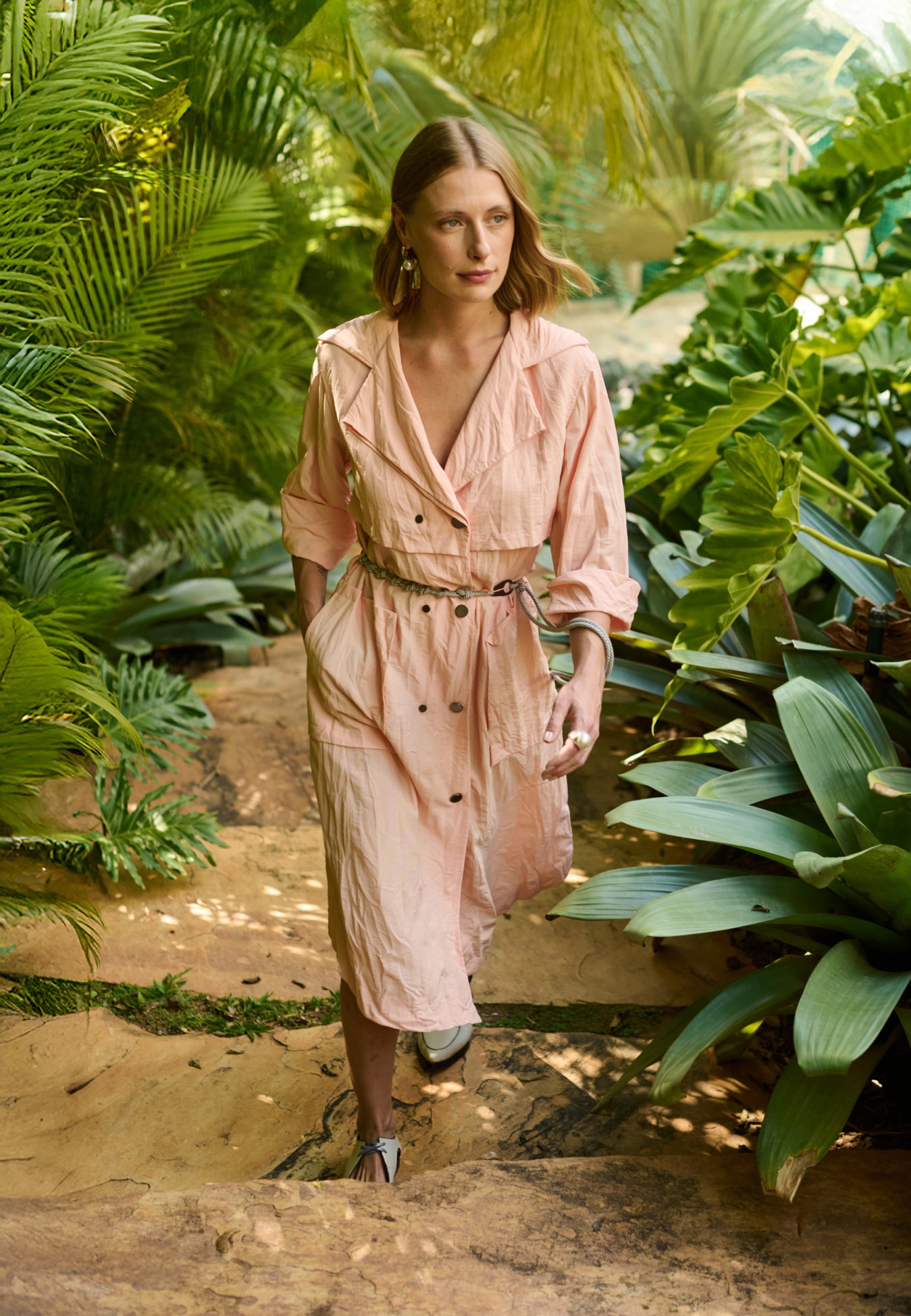 Modelo veste vestido midi na cor pêssego em tecido leve. A peça possui fechamento transpassado com botões metálicos, gola de alfaiataria, bolsos frontais grandes e cinto de cordão náutico.