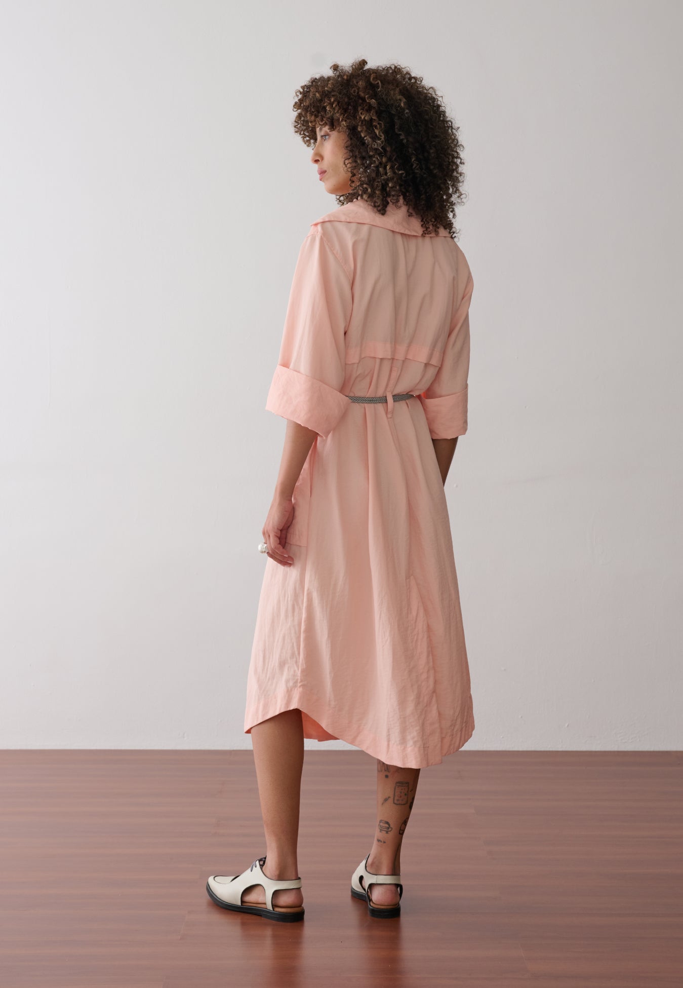 Modelo de costas veste vestido midi na cor pêssego em tecido leve. A peça possui recorte horizontal e cinto de cordão náutico.