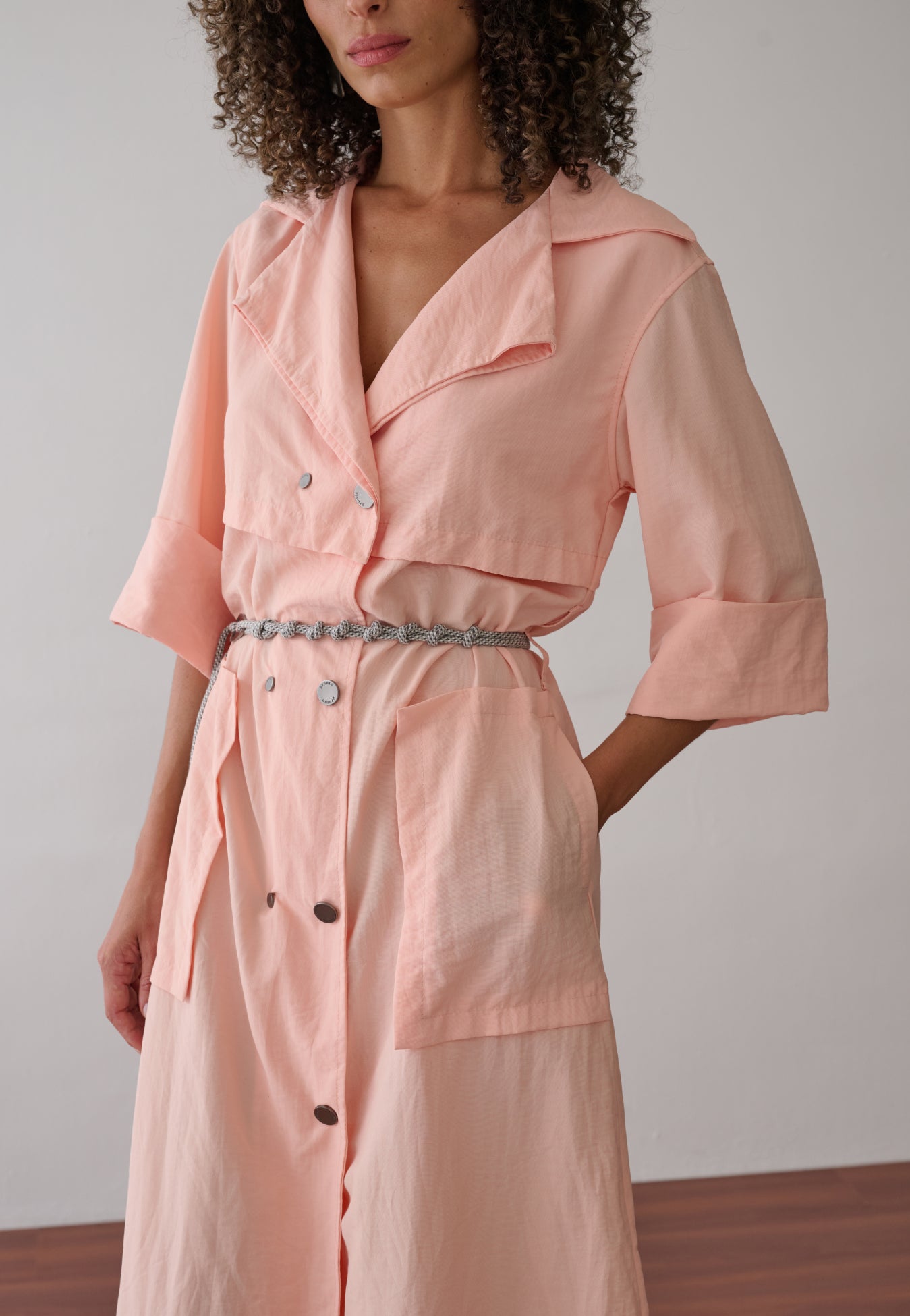 Modelo veste vestido midi na cor pêssego em tecido leve. A peça possui fechamento transpassado com botões metálicos, gola de alfaiataria, bolsos frontais grandes e cinto de cordão náutico.