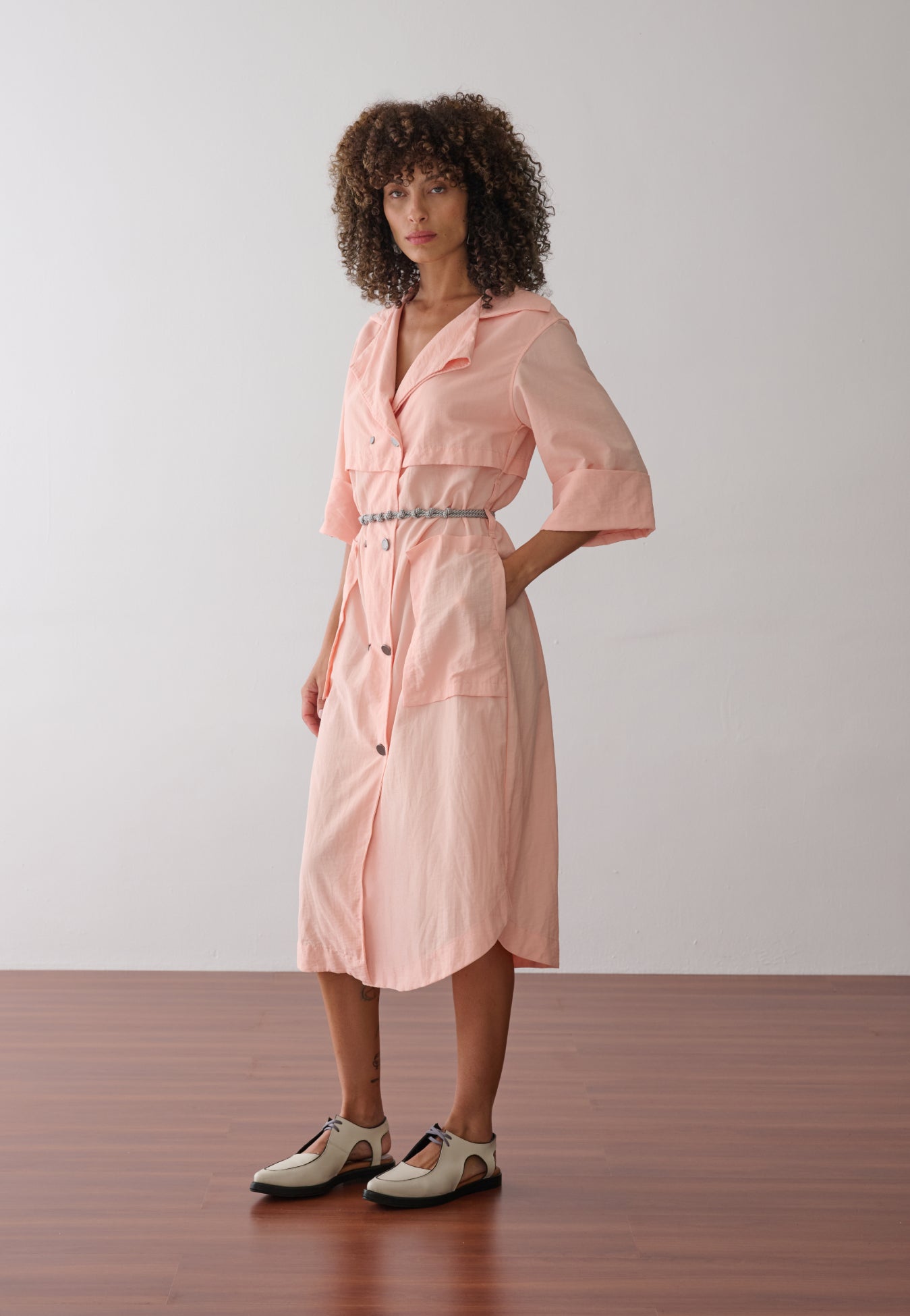 Modelo veste vestido midi na cor pêssego em tecido leve. A peça possui fechamento transpassado com botões metálicos, gola de alfaiataria, bolsos frontais grandes e cinto de cordão náutico.