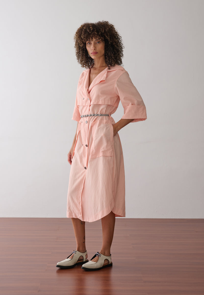 Modelo veste vestido midi na cor pêssego em tecido leve. A peça possui fechamento transpassado com botões metálicos, gola de alfaiataria, bolsos frontais grandes e cinto de cordão náutico.