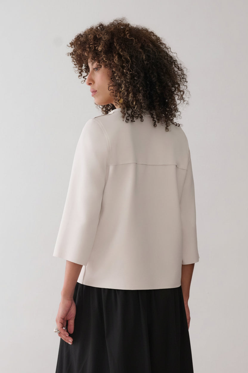 Modelo de costas usando uma blusa off-white de mangas 3/4 com costura horizontal aparente, combinada com uma saia preta de tecido leve e fluido.