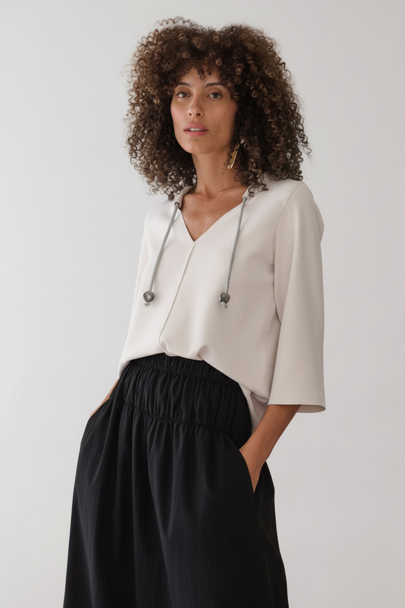 Modelo veste uma blusa off-white com mangas 3/4 e decote em "V" com cordões decorativos, combinada com uma saia midi preta de cós elástico franzido.
