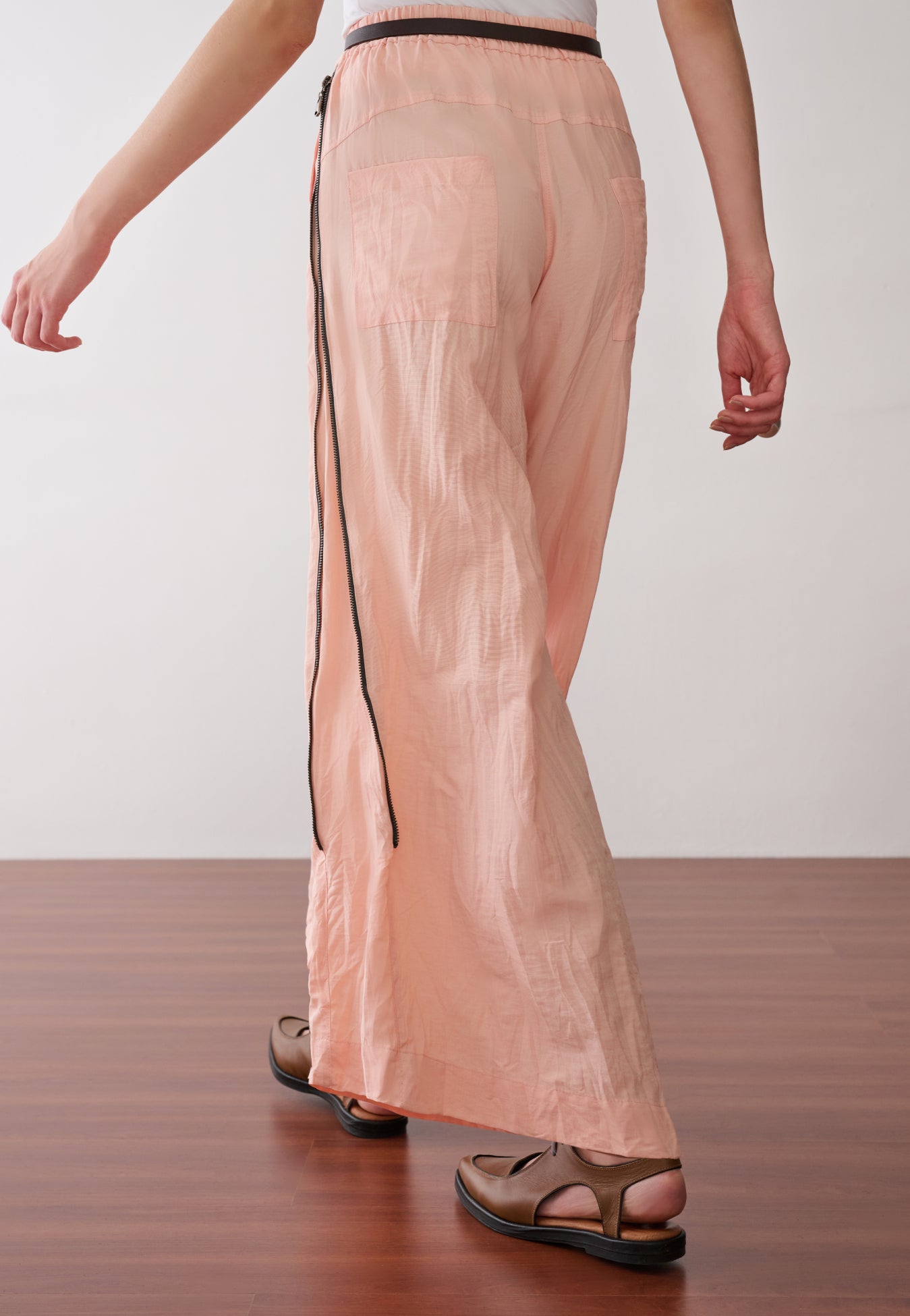 Modelo de costas veste uma calça pantalona na cor pêssego, com cós elástico, bolsos aplicados e zíper nas laterais, cinto fino e sapatos marrons de bico fino.