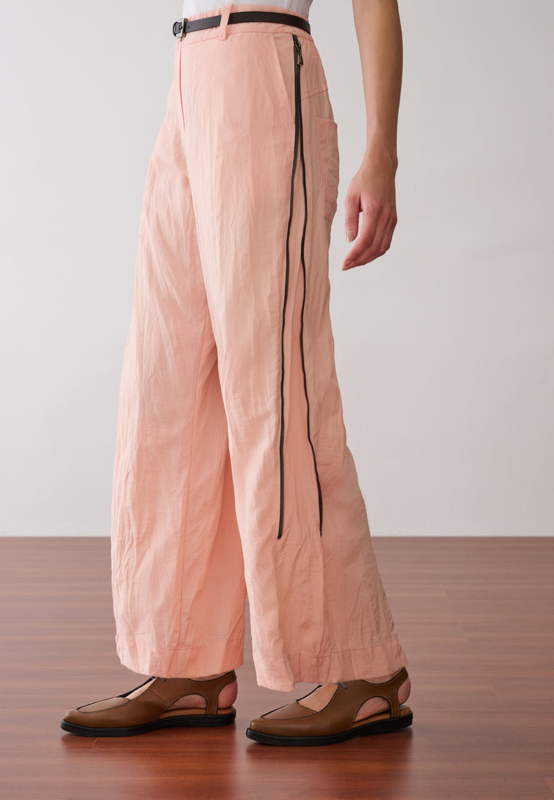 Modelo veste uma calça pantalona na cor pêssego com zíper nas laterais, cinto fino marrom com fivela de metal e sapatos marrons de bico fino.