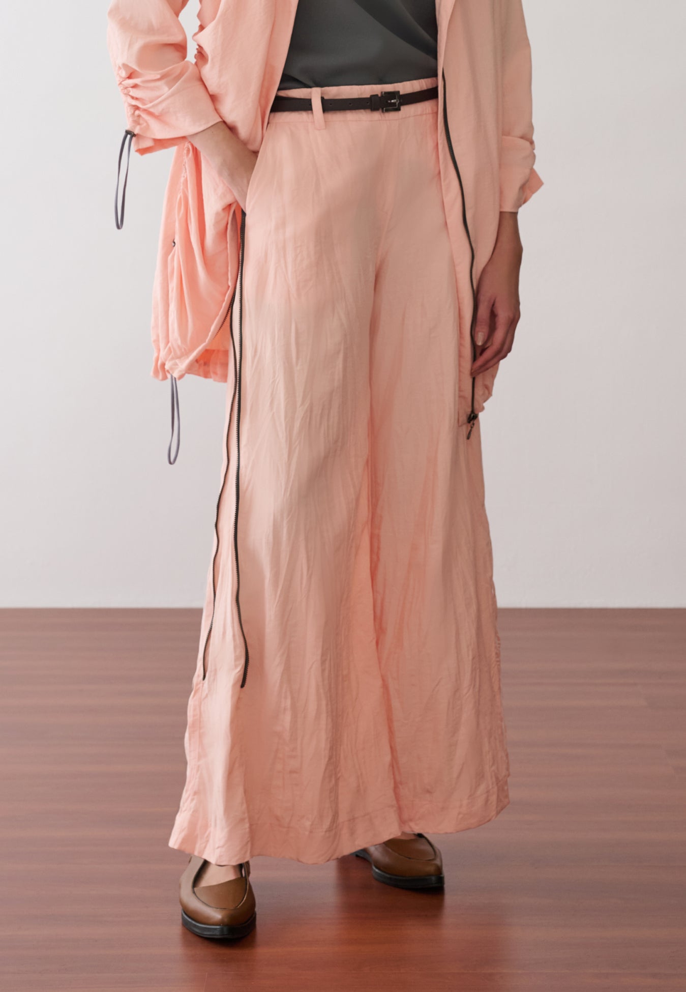 Modelo veste uma calça pantalona na cor pêssego com zíper nas laterais, cinto fino marrom com fivela de metal e sapatos marrons de bico fino.