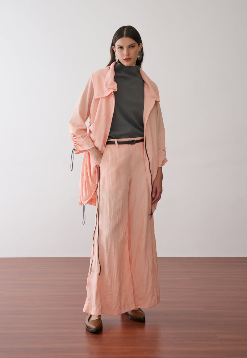 Modelo veste uma jaqueta longa com capuz e cordões de ajuste na cor pêssego sobre uma blusa cinza de gola alta, combinada com calças pantalona pêssego com zíper nas laterais, cinto fino marrom com fivela de metal e sapatos marrons de bico fino.