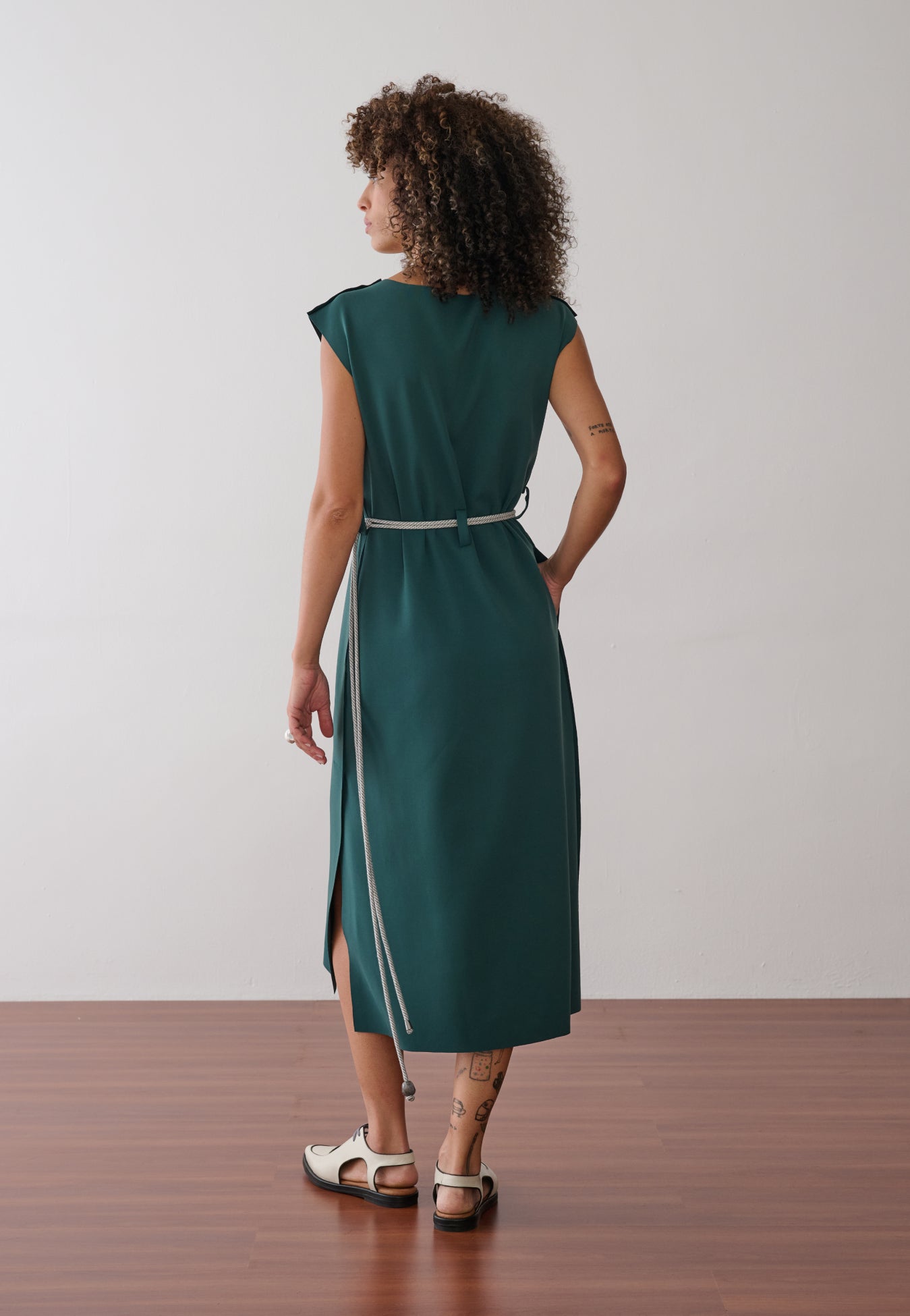 Modelo de costas exibe um vestido midi verde-petróleo sem mangas, com fenda lateral, acinturado por um cordão bicolor fino e combinado com sapatos off-white de bico fino e recortes.