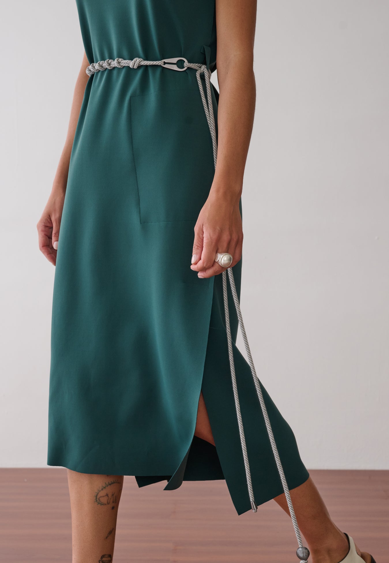 Modelo usa um vestido midi verde-petróleo sem mangas, com fenda lateral, acinturado por um cordão bicolor fino e combinado com sapatos off-white de bico fino e recortes.