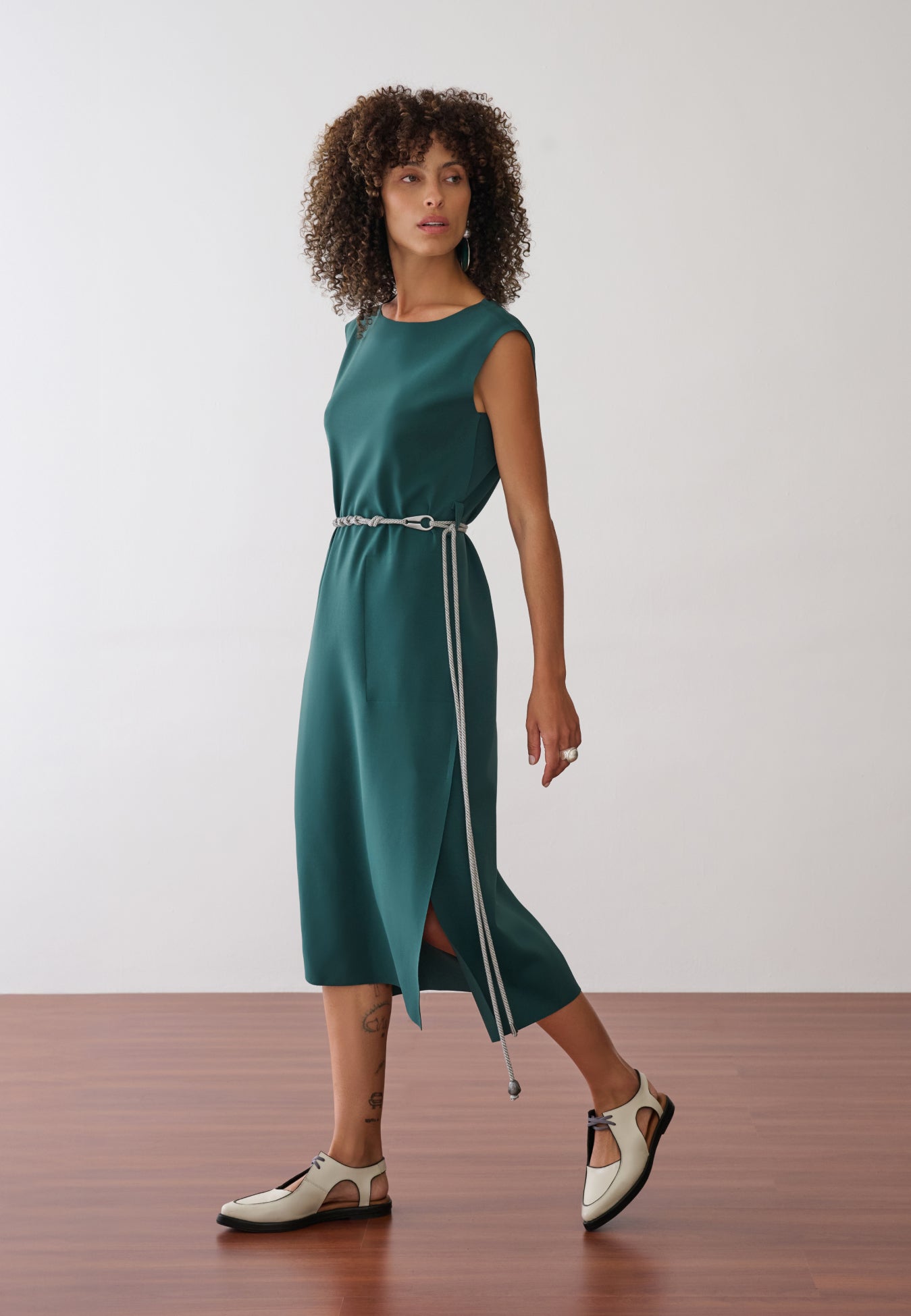 Modelo usa um vestido midi verde-petróleo sem mangas, com fenda lateral, acinturado por um cordão bicolor fino e combinado com sapatos off-white de bico fino e recortes.