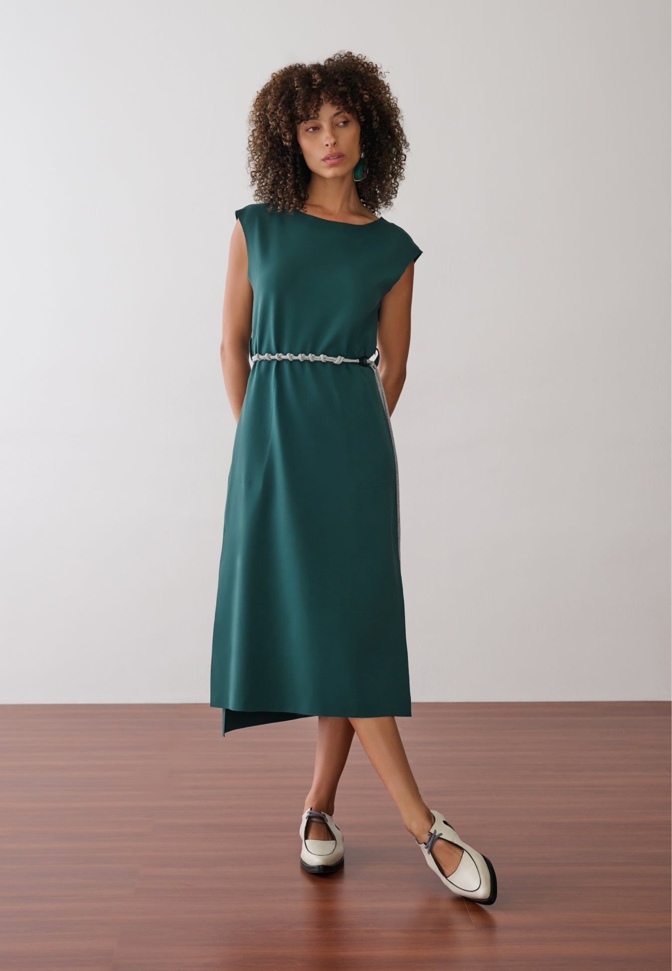 Modelo usa um vestido midi verde-petróleo sem mangas, com fenda lateral, acinturado por um cordão bicolor fino e combinado com sapatos off-white de bico fino e recortes.