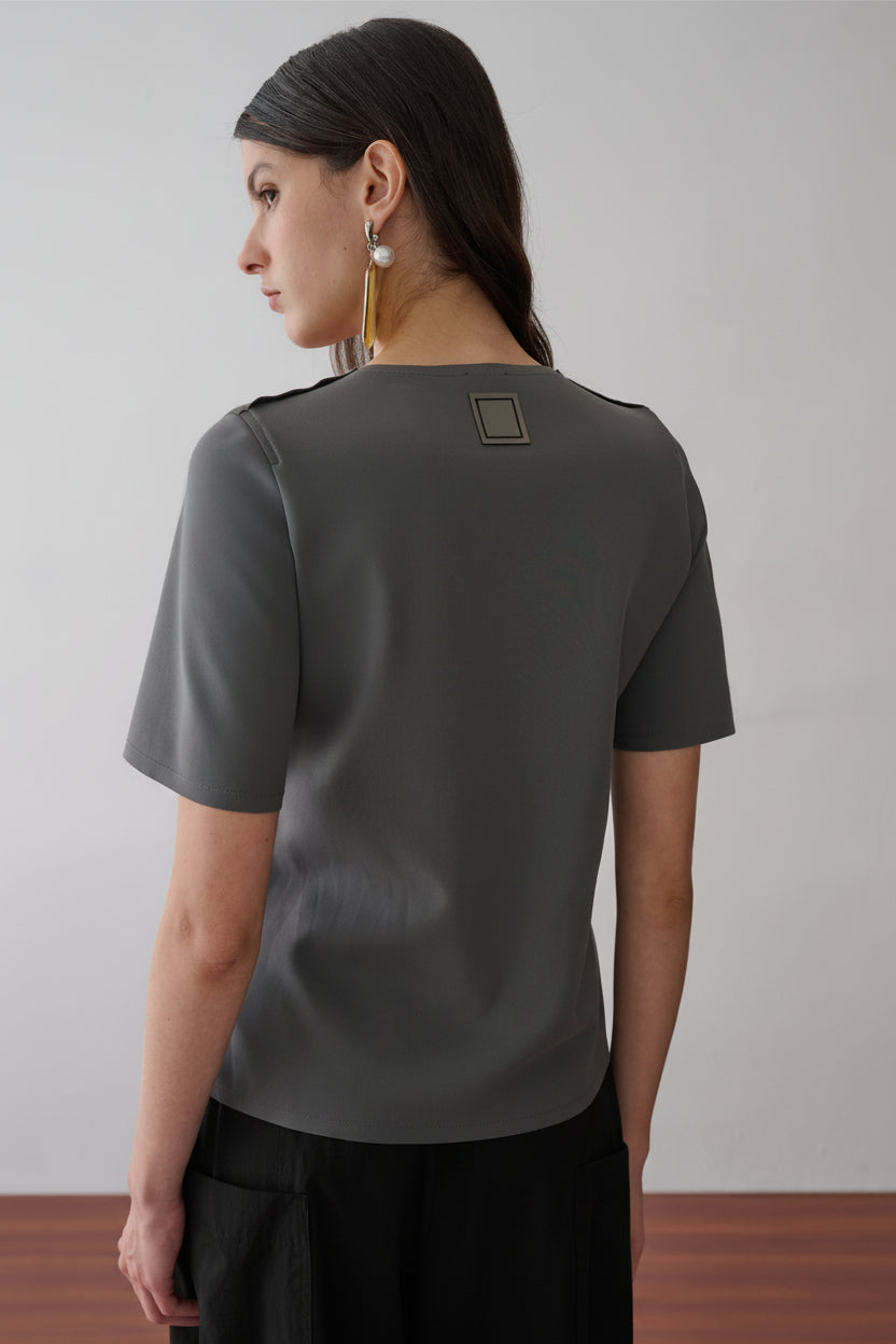Modelo de costas usando uma blusa cinza de mangas curtas com uma etiqueta retangular decorativa centralizada na parte superior, combinada com uma saia preta.