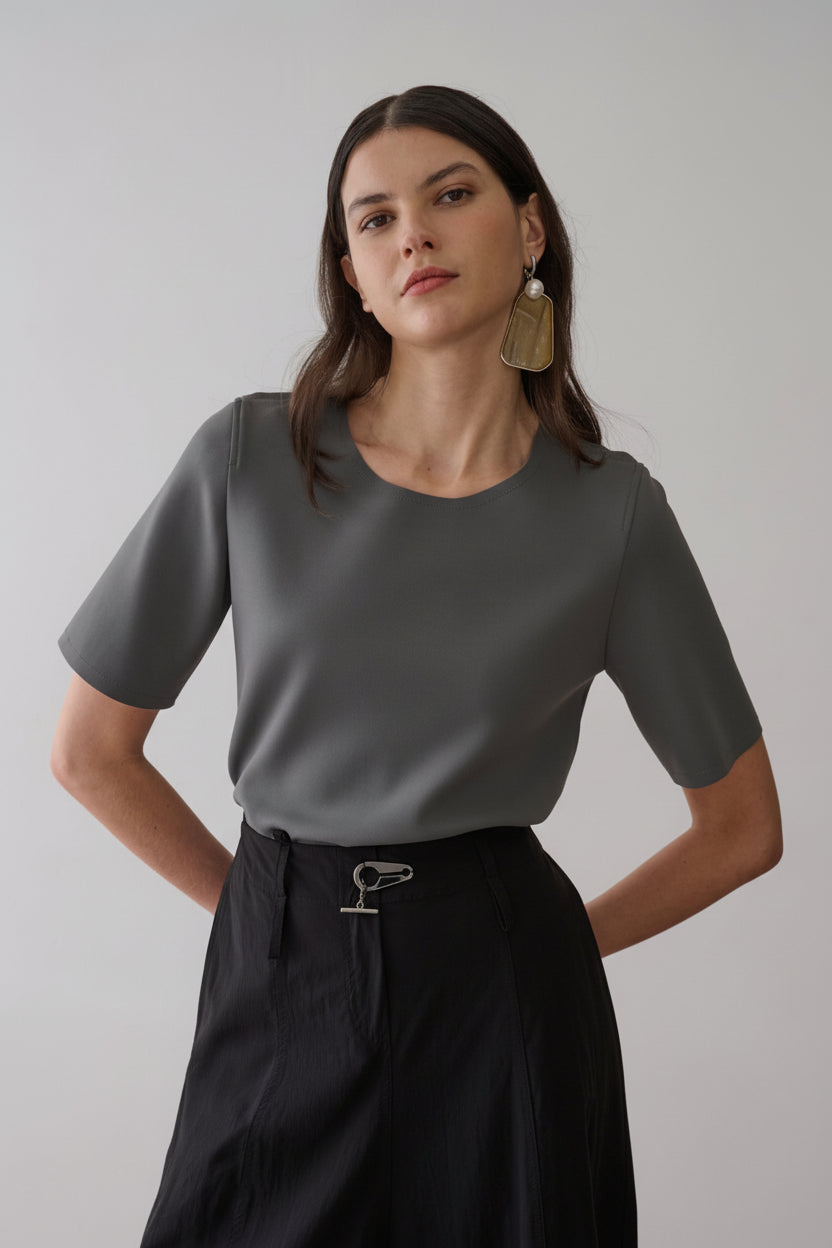 Modelo veste uma blusa cinza de decote arredondado e mangas curtas, combinada com uma saia preta que apresenta um fecho metálico diferenciado no cós.