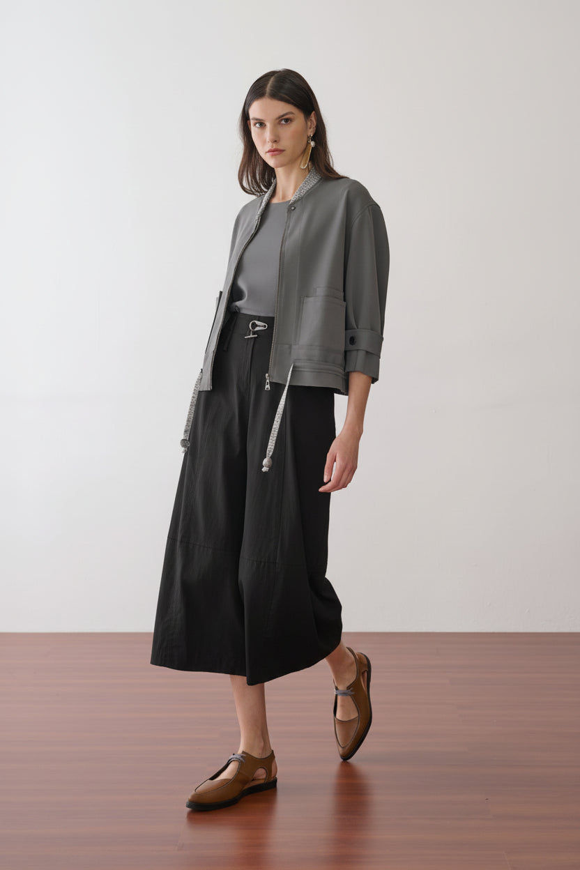 Modelo veste uma jaqueta cinza de mangas 3/4 com abotoamento nos punhos sobre uma blusa cinza, combinada com calça preta volumosa, cordão prateado na cintura e sapatos marrons de bico fino.