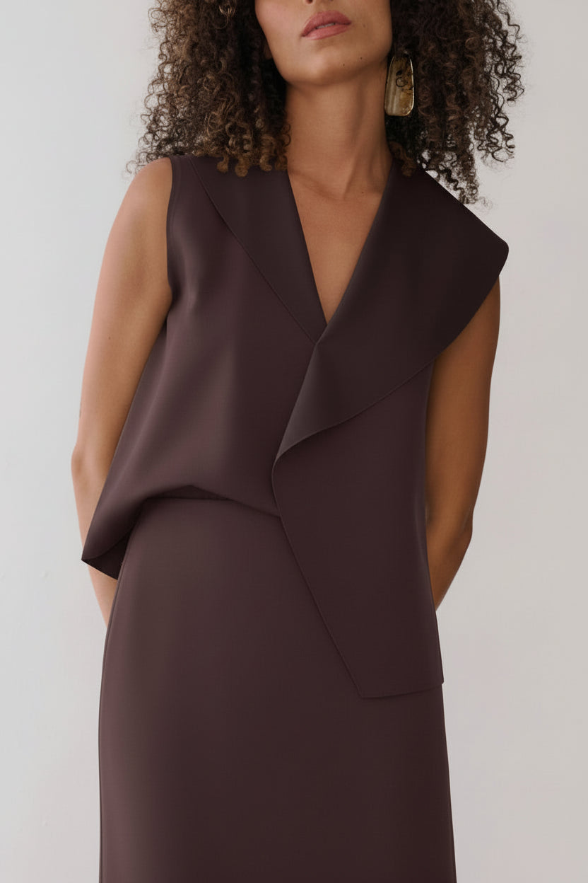 Modelo veste uma blusa marrom sem mangas com lapelas assimétricas e gola em "V", combinada com saia reta da mesma cor.