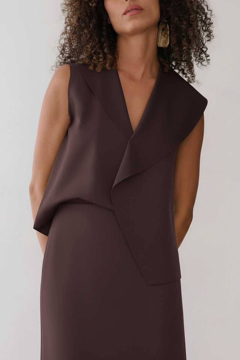 Modelo veste uma blusa marrom sem mangas com lapelas assimétricas e gola em "V", combinada com saia reta da mesma cor.