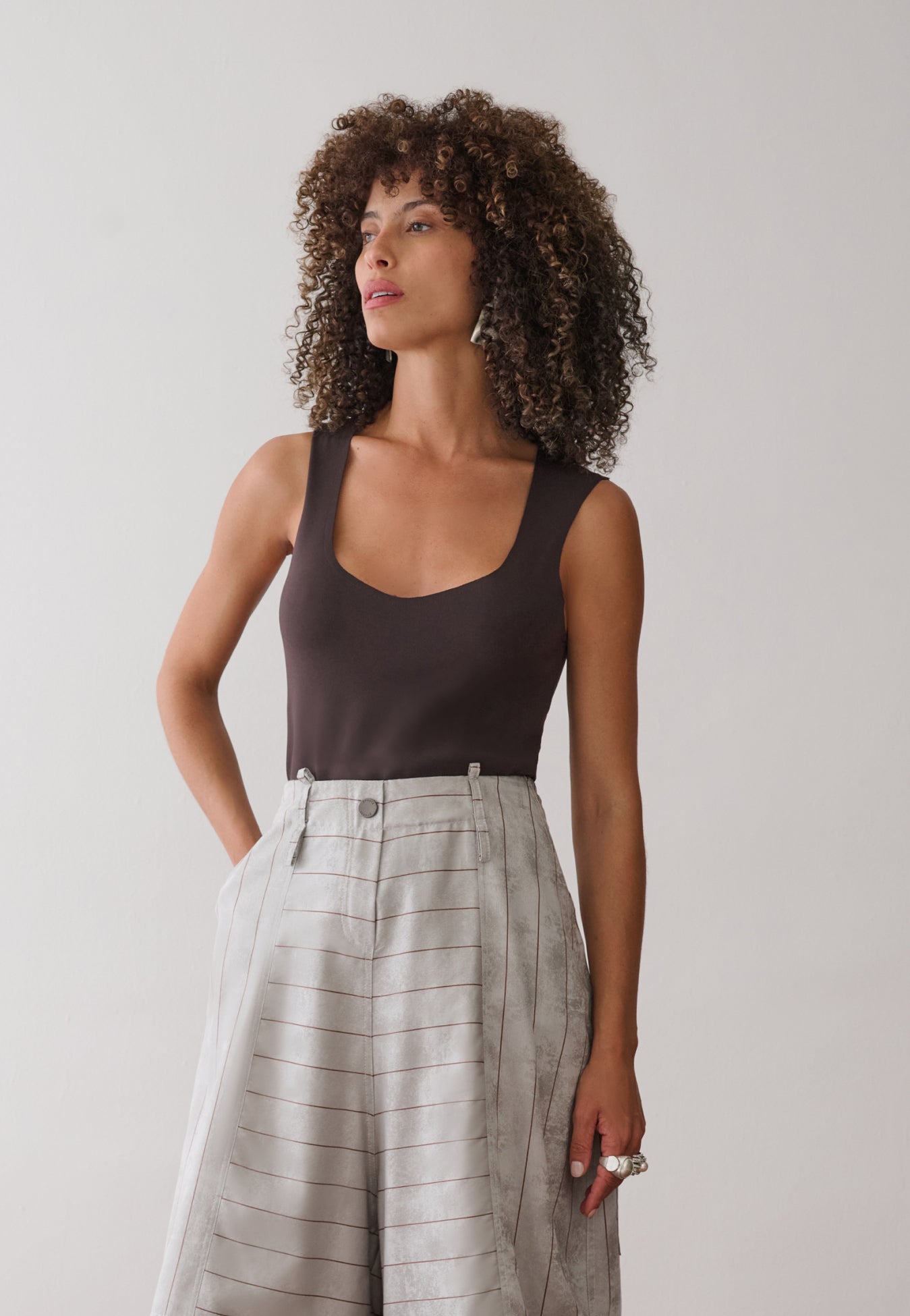 Modelo veste uma regata marrom de decote arredondado combinada com calça pantacourt cinza com listras marrons de cintura alta e bolsos laterais.