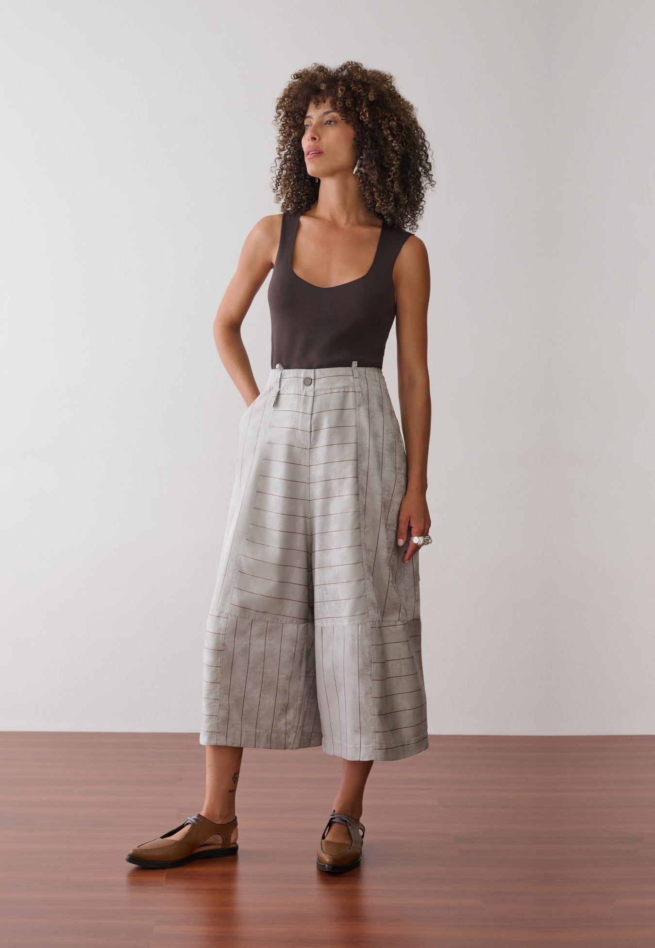 Modelo veste uma regata marrom de decote arredondado combinada com calças pantacourt cinza com listras marrons de cintura alta, bolsos laterais e sapatos marrons de bico fino. 