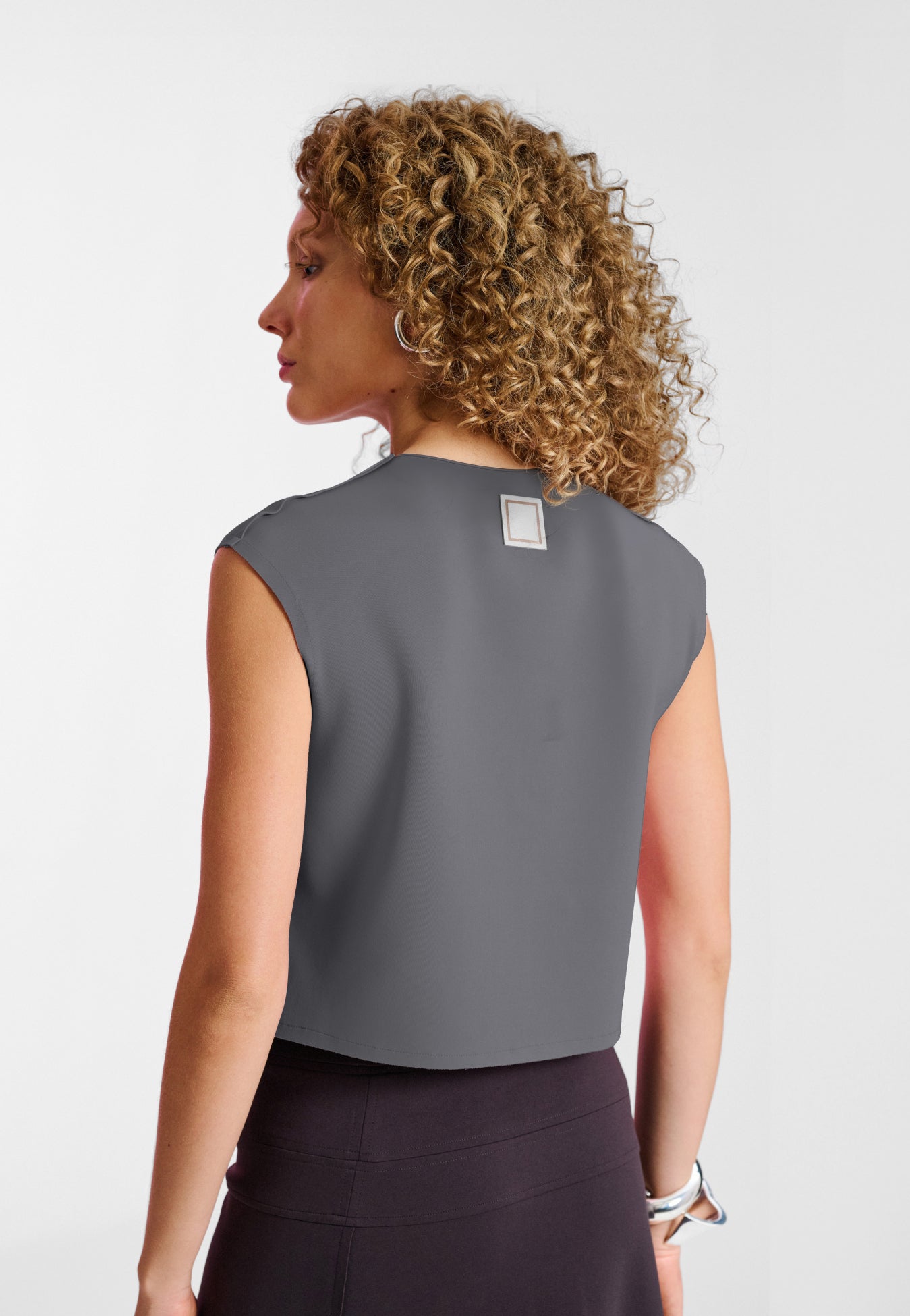 Modelo de costas usando uma regata cinza ajustada com uma pequena etiqueta quadrada no decote, combinada com calça marrom.
