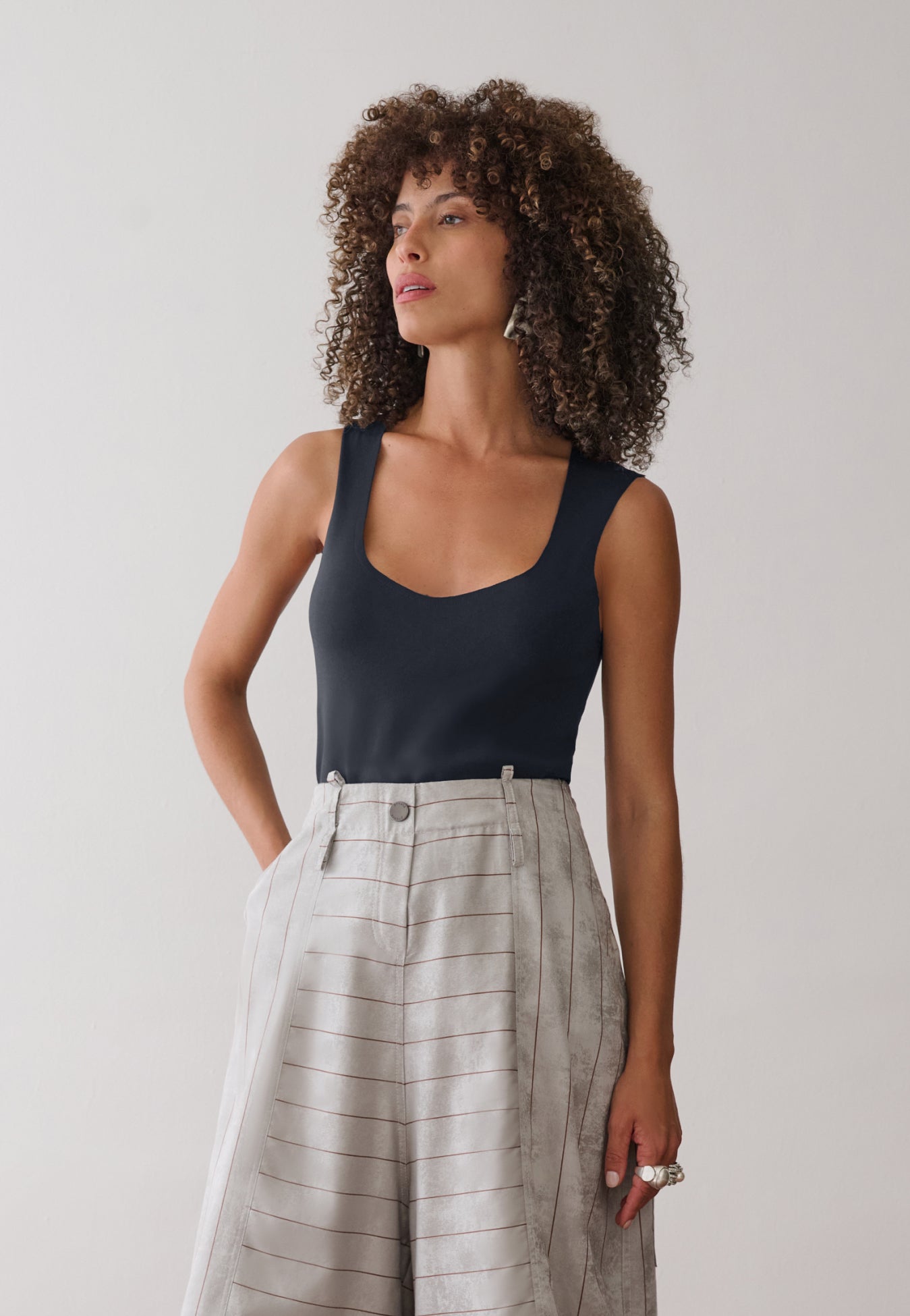 Modelo veste uma regata preta de decote arredondado combinada com calça pantacourt cinza com listras marrons de cintura alta e bolsos laterais.