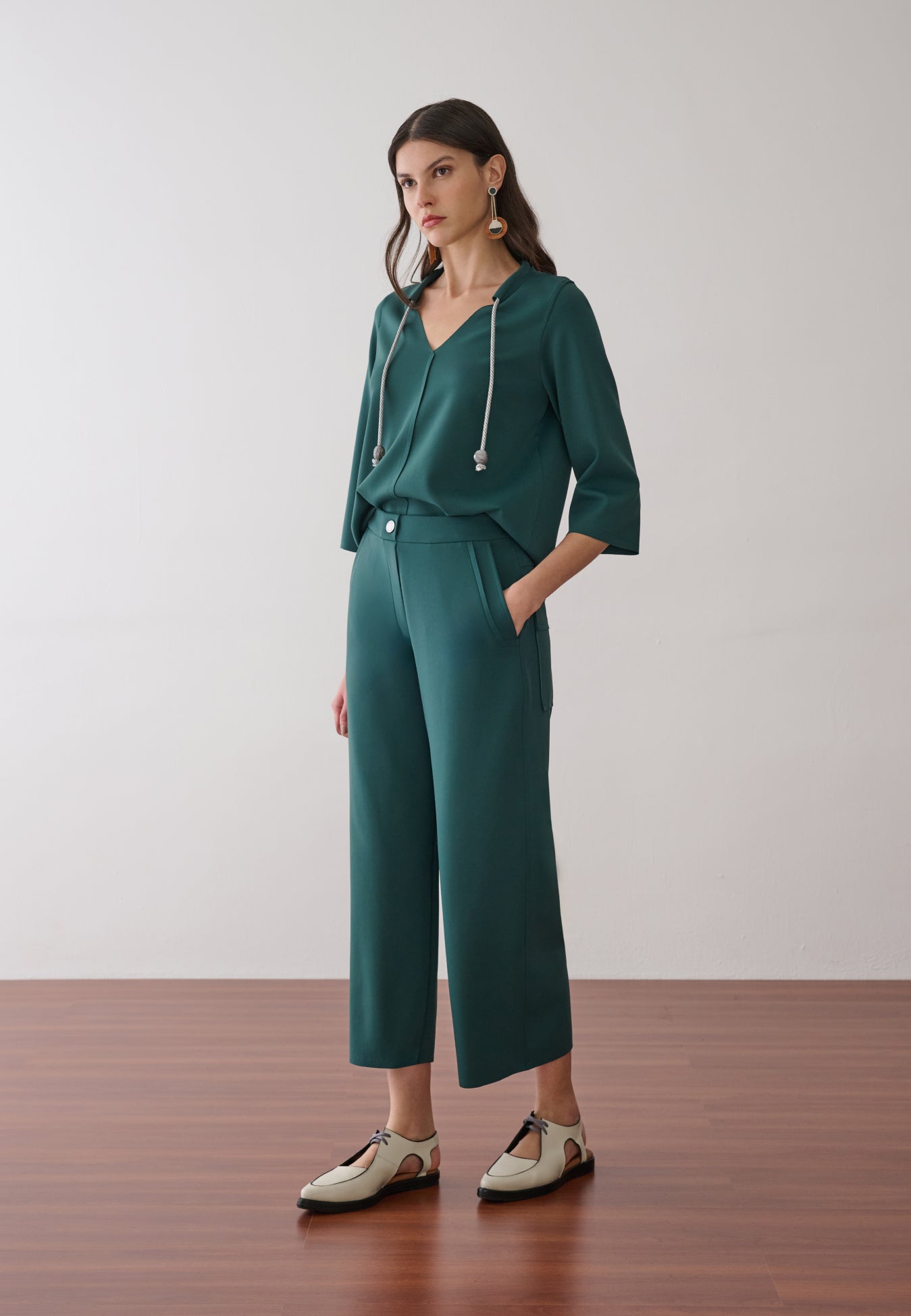 Modelo veste um conjunto verde-petróleo composto por blusa de decote em "V" com cordões decorativos e calça pantacourt de alfaiataria, finalizado com sapatos off-white de bico fino e recortes.