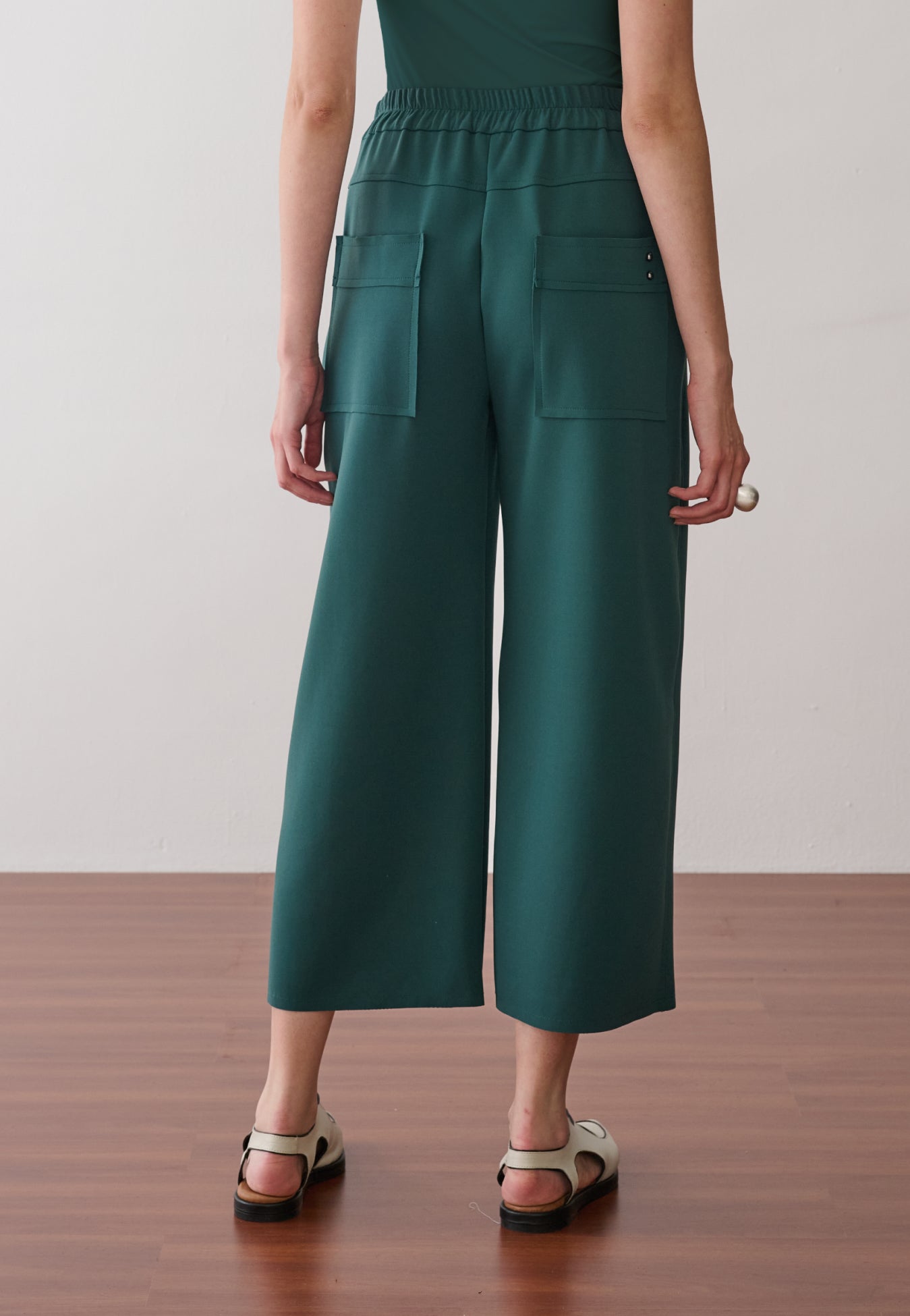 Modelo de costas usando uma calça pantacourt verde-petróleo com cós elástico, bolsos traseiros quadrados e amplos e sapatos off-white com recortes.