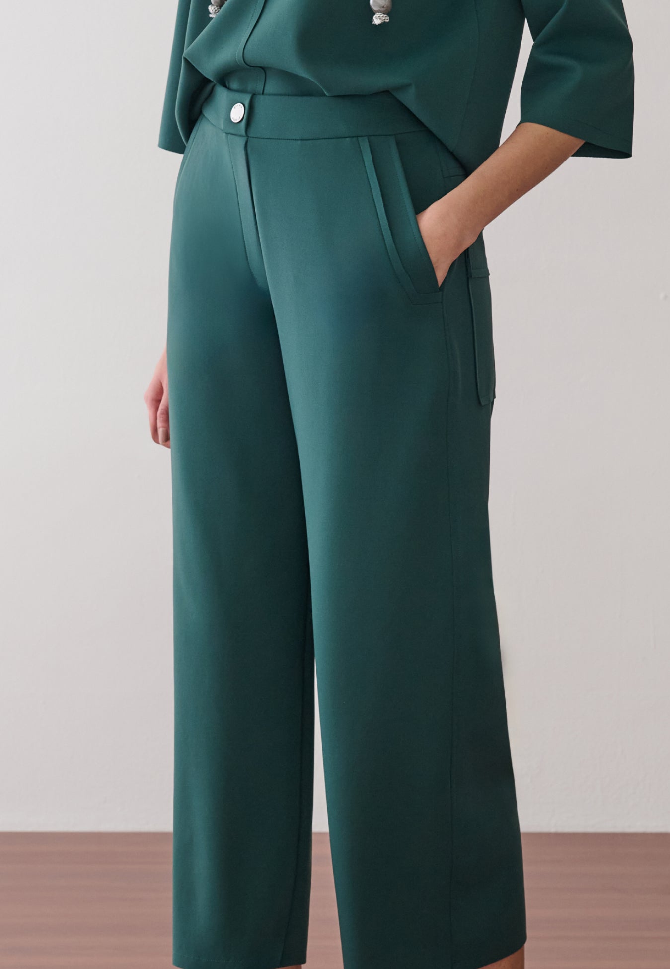 Modelo veste calça pantacourt verde-petróleo, destacando o bolso faca com acabamento pespontado, o cós com botão metálico central e o caimento estruturado do tecido.
