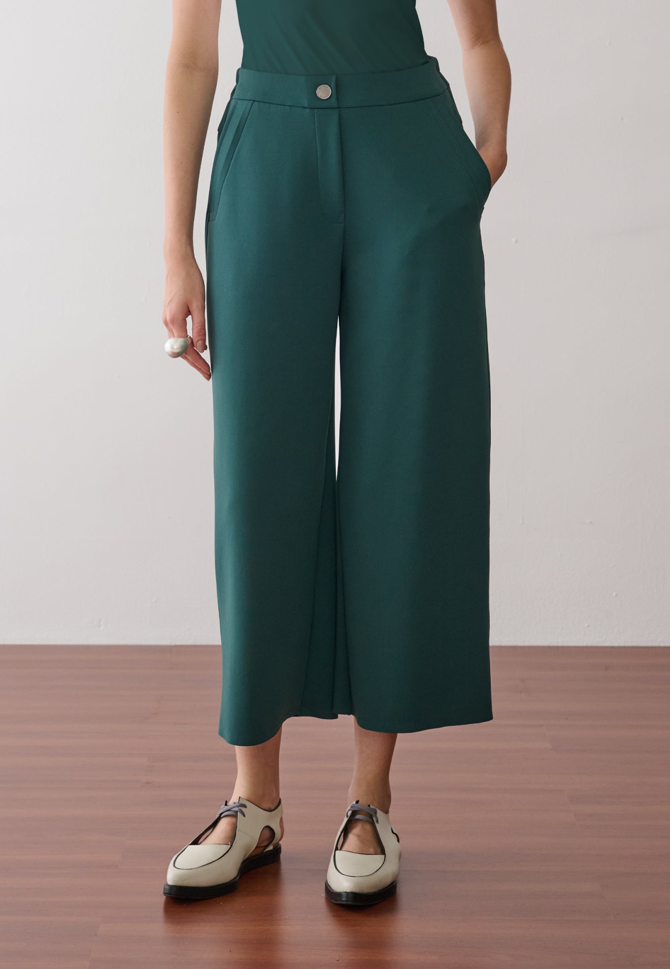Modelo veste calça pantacourt verde-petróleo, destacando o bolso faca com acabamento pespontado, o cós com botão metálico central e o caimento estruturado do tecido, finalizado com sapatos off-white de bico fino e recortes.