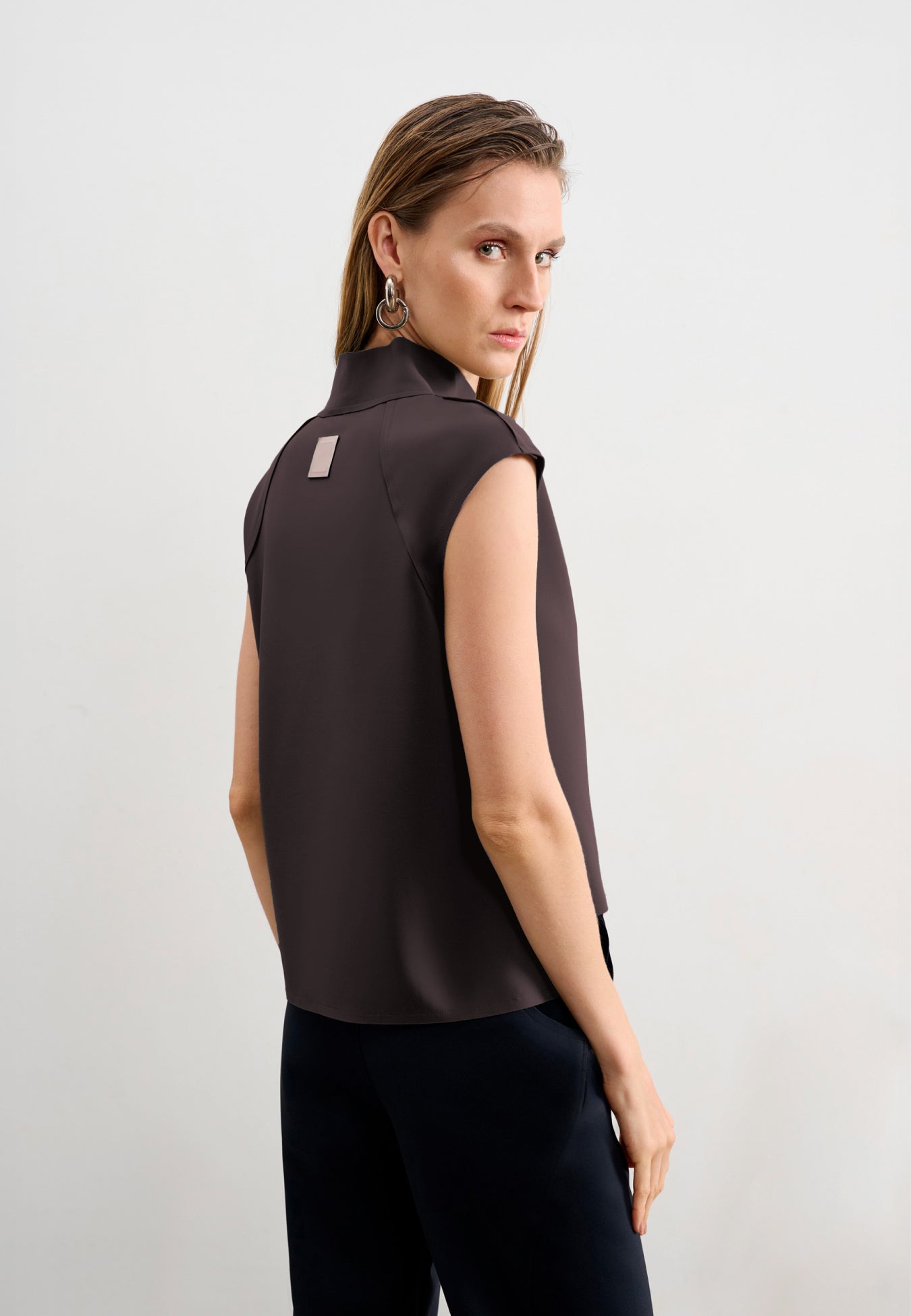 Modelo de costas usando uma blusa marrom sem mangas de gola alta com uma etiqueta retangular decorativa centralizada na parte superior, combinada com calça preta.