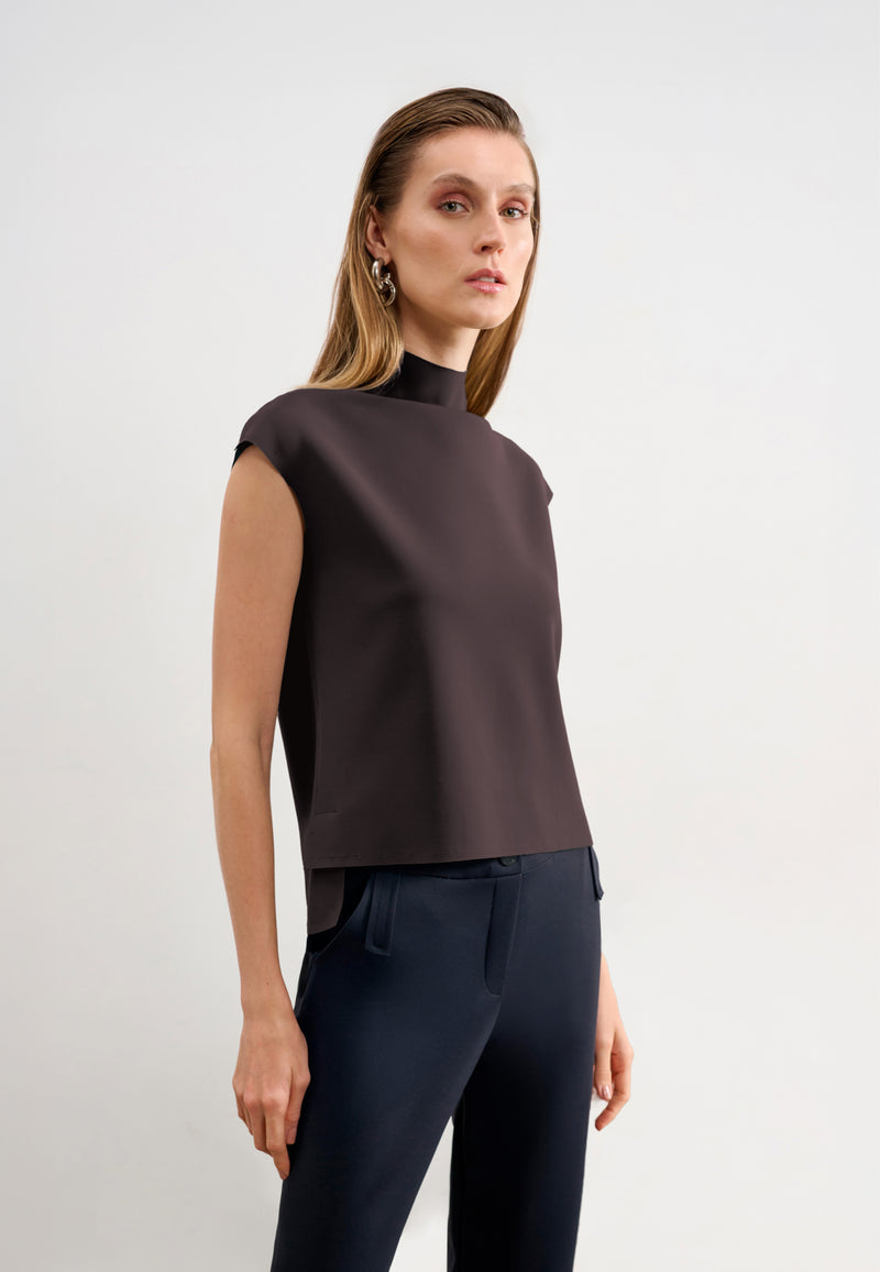 Modelo veste uma blusa marrom sem mangas de gola alta com barra assimétrica, combinada com calças pretas.