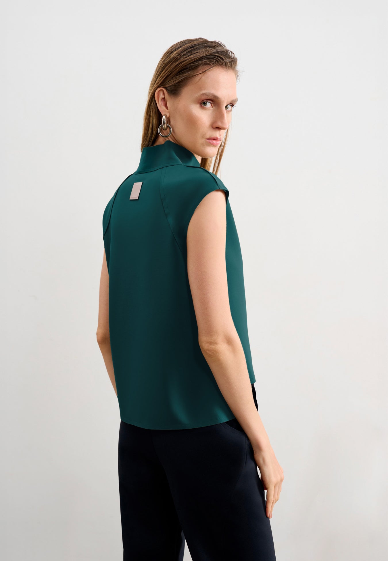 Modelo de costas usando uma blusa verde-petróleo sem mangas de gola alta com uma etiqueta retangular decorativa centralizada na parte superior, combinada com calça branca.