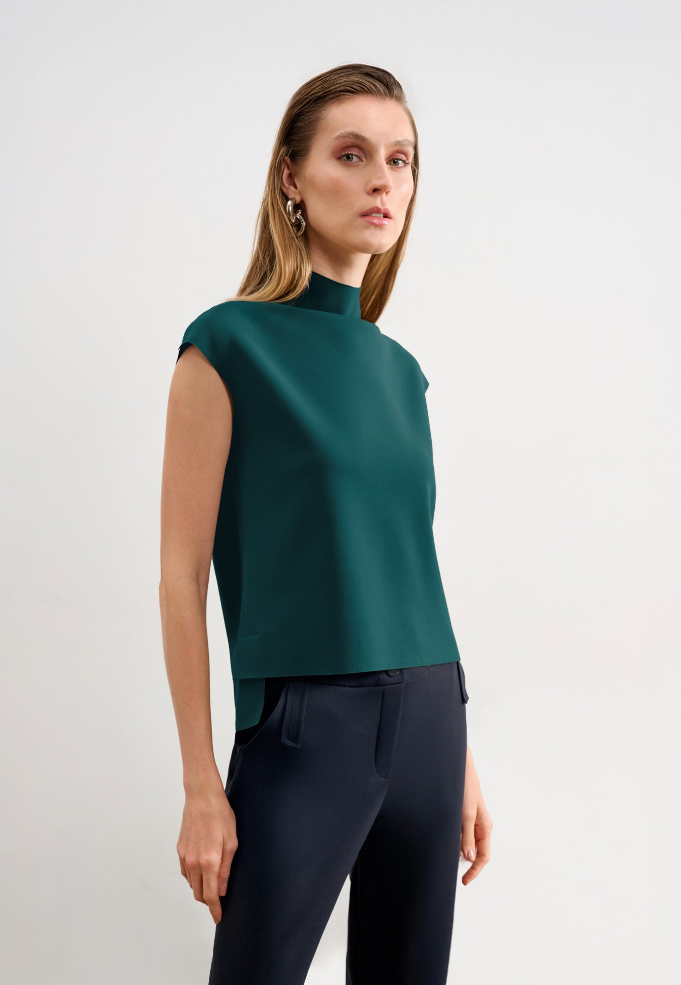 Modelo veste uma blusa verde-petróleo sem mangas de gola alta com barra assimétrica, combinada com calças pretas.