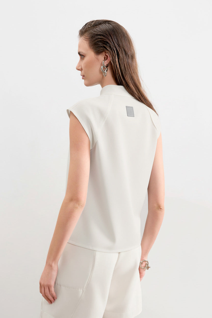Modelo de costas usando uma blusa branca sem mangas de gola alta com uma etiqueta retangular decorativa centralizada na parte superior, combinada com calça de mesma cor.