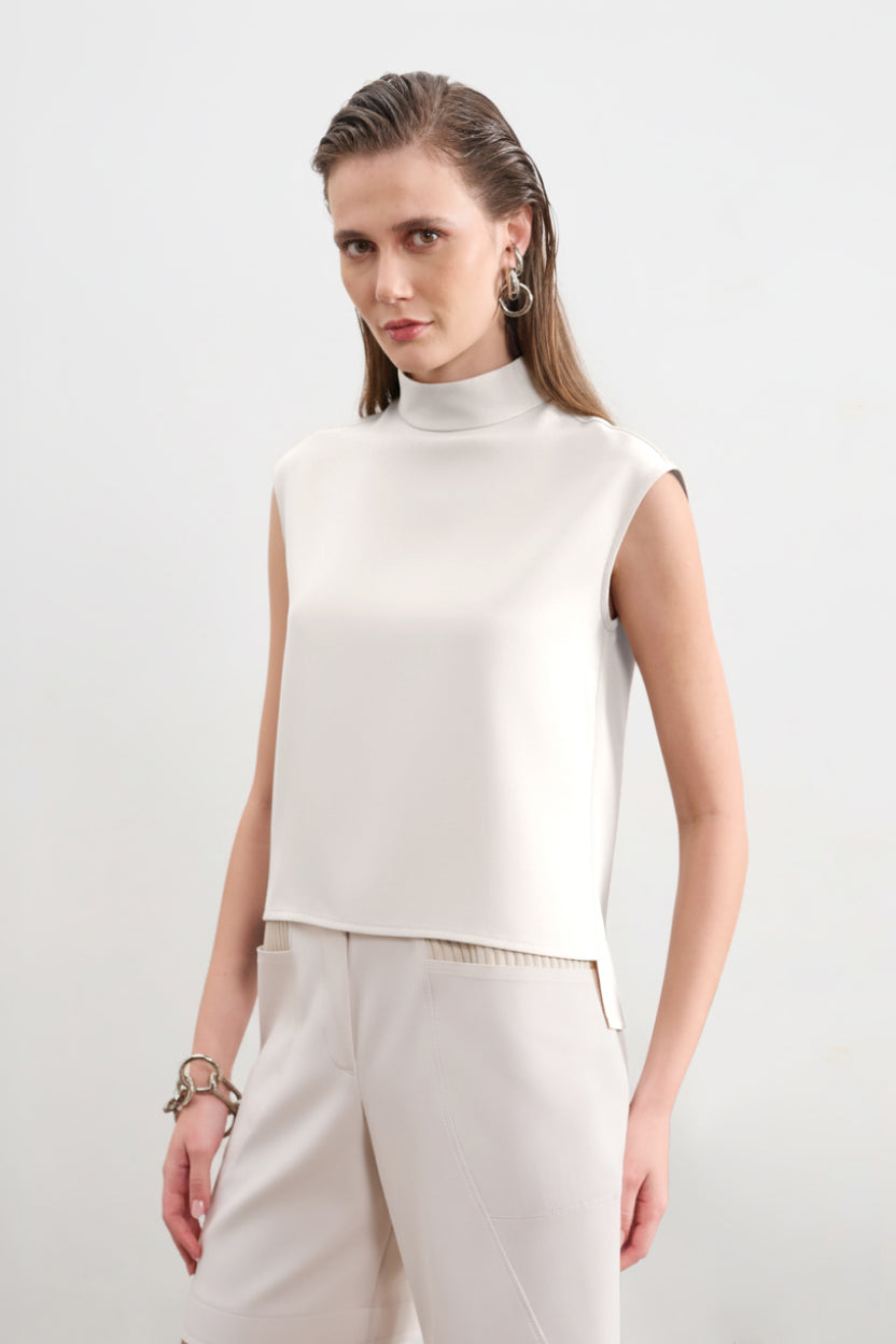 Modelo veste uma blusa branca sem mangas de gola alta com barra assimétrica, combinada com calças da mesma cor. 