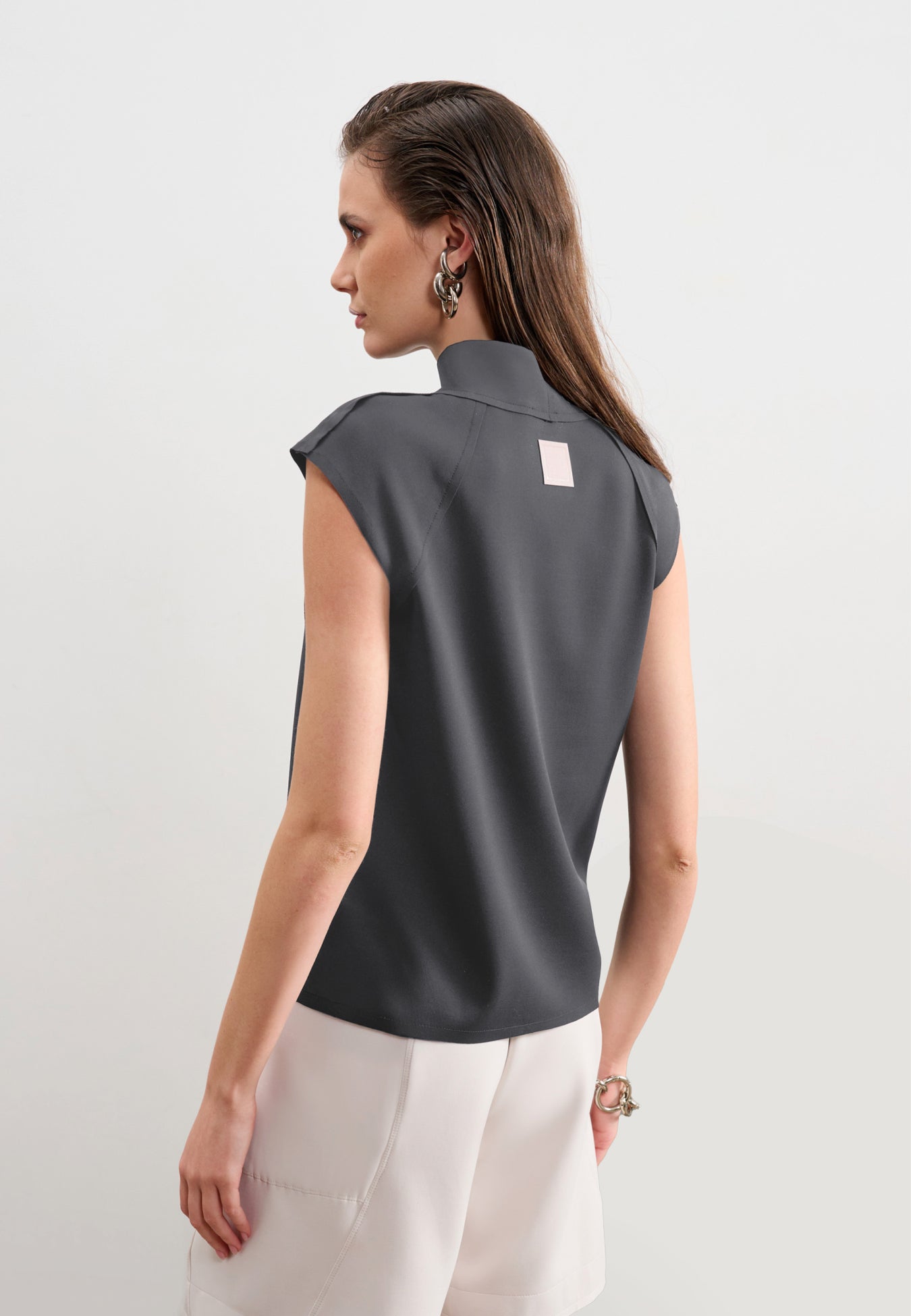 Modelo de costas usando uma blusa cinza sem mangas de gola alta com uma etiqueta retangular decorativa centralizada na parte superior, combinada com calça branca.