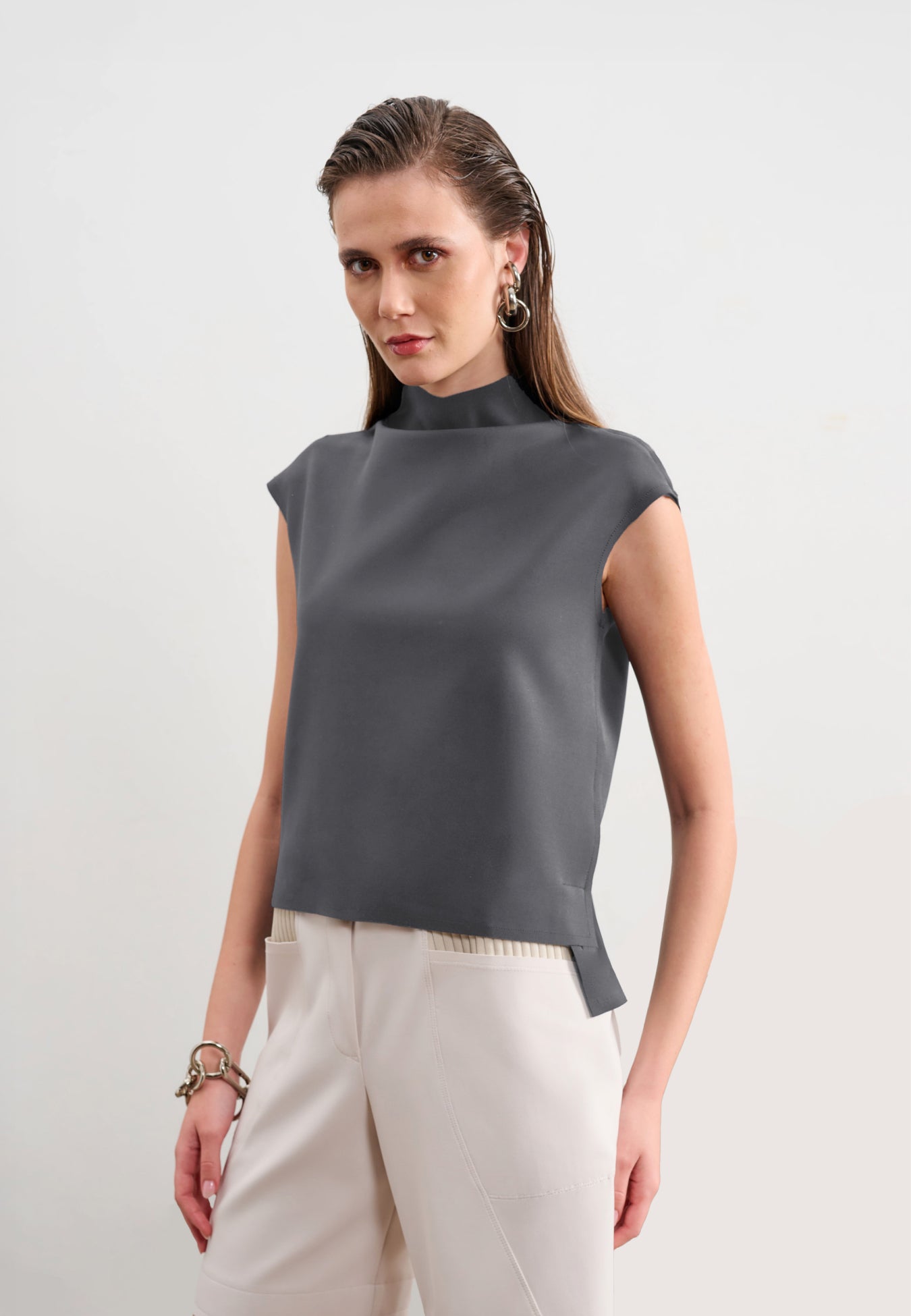 Modelo veste uma blusa cinza sem mangas de gola alta com barra assimétrica, combinada com calças brancas.