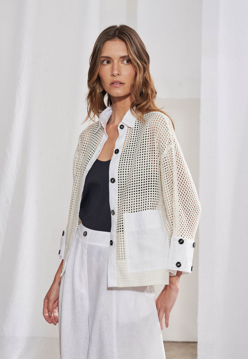 Mulher vestindo camisa de modelagem boxy em malha telada com transparência sutil. Gola colarinho, vista frontal com botões pretos, punhos largos e barra em tecido plano liso marfim. Bolso quadrado chapado no lado esquerdo. A camisa está aberta como terceira peça sobre regata preta e calça branca solta.