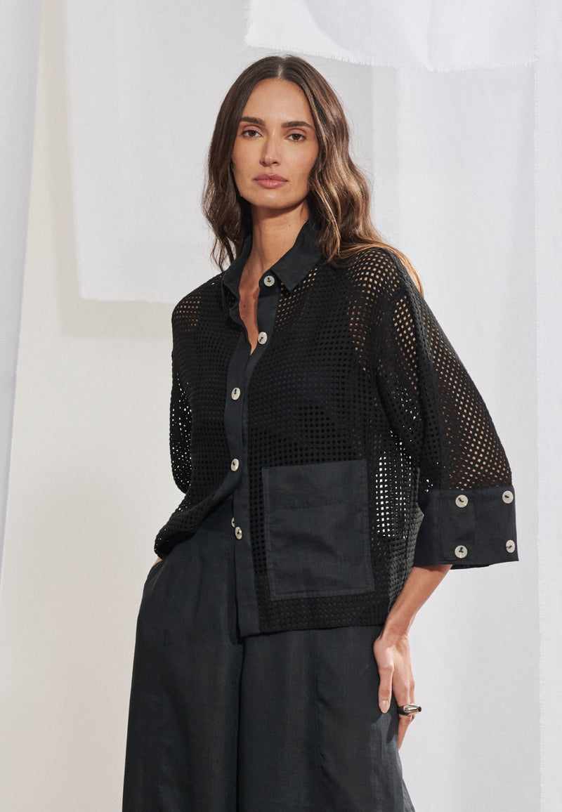 Mulher vestindo camisa de modelagem boxy em malha telada com transparência sutil. Gola colarinho, vista frontal com botões pretos, punhos largos e barra em tecido plano liso marfim. Bolso quadrado chapado no lado esquerdo. A camisa está aberta como terceira peça sobre regata preta e calça branca solta.