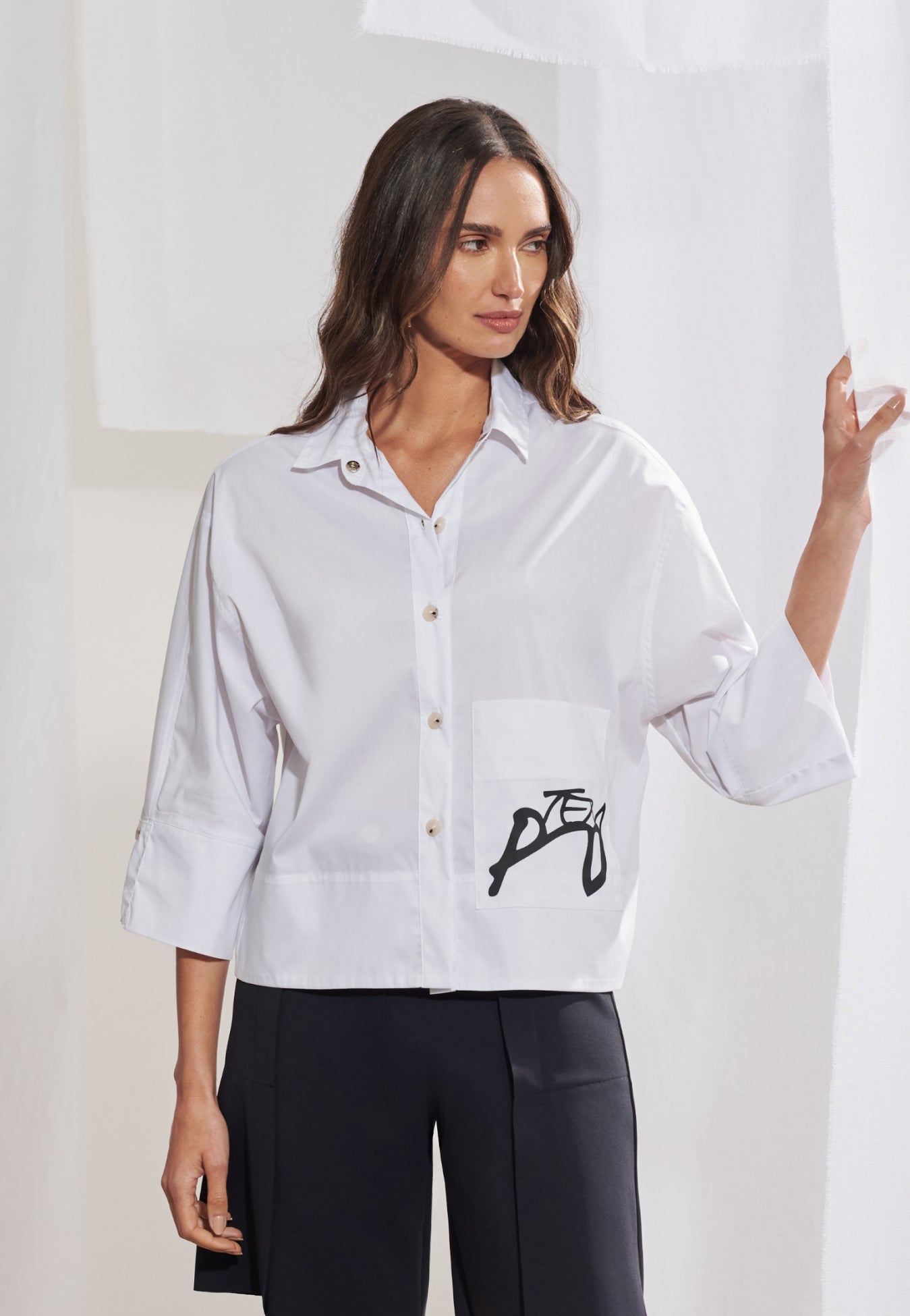 Mulher vestindo camisa boxy em tricoline de modelagem ampla com comprimento no quadril. A peça tem gola colarinho clássica, mangas 3/4 com punhos largos e botões escuros contrastantes. Bolso frontal esquerdo com estampa geométrica preta localizada. Combinada com bermuda de alfaiataria preta de corte reto.