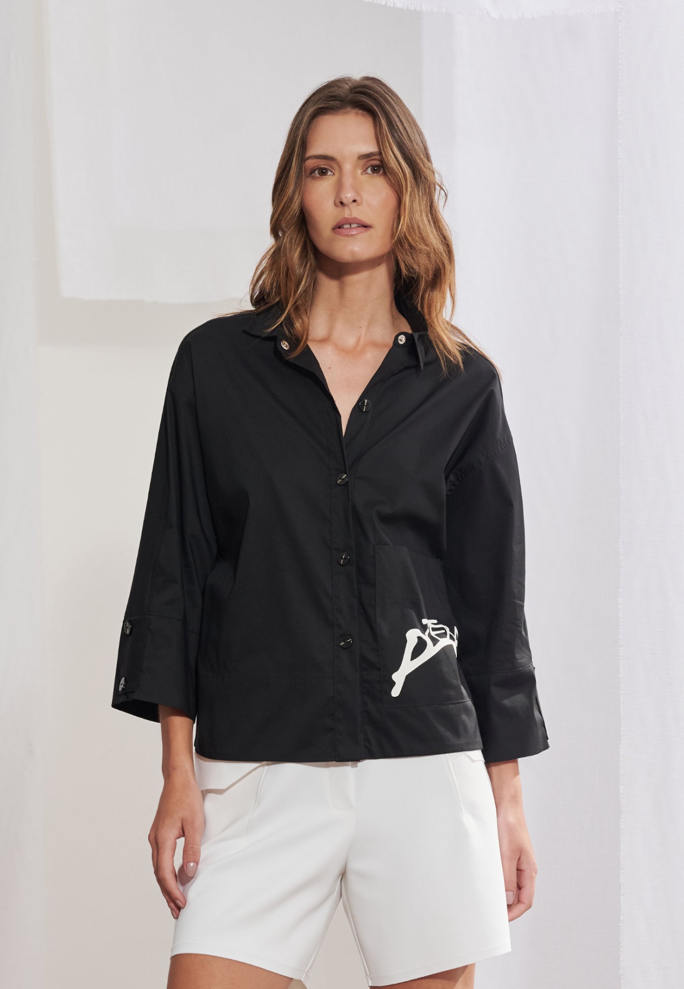 Mulher vestindo camisa boxy em tricoline de modelagem ampla com comprimento no quadril. A peça tem gola colarinho clássica, mangas 3/4 com punhos largos e botões escuros contrastantes. Bolso frontal esquerdo com estampa geométrica preta localizada. Combinada com bermuda de alfaiataria preta de corte reto.